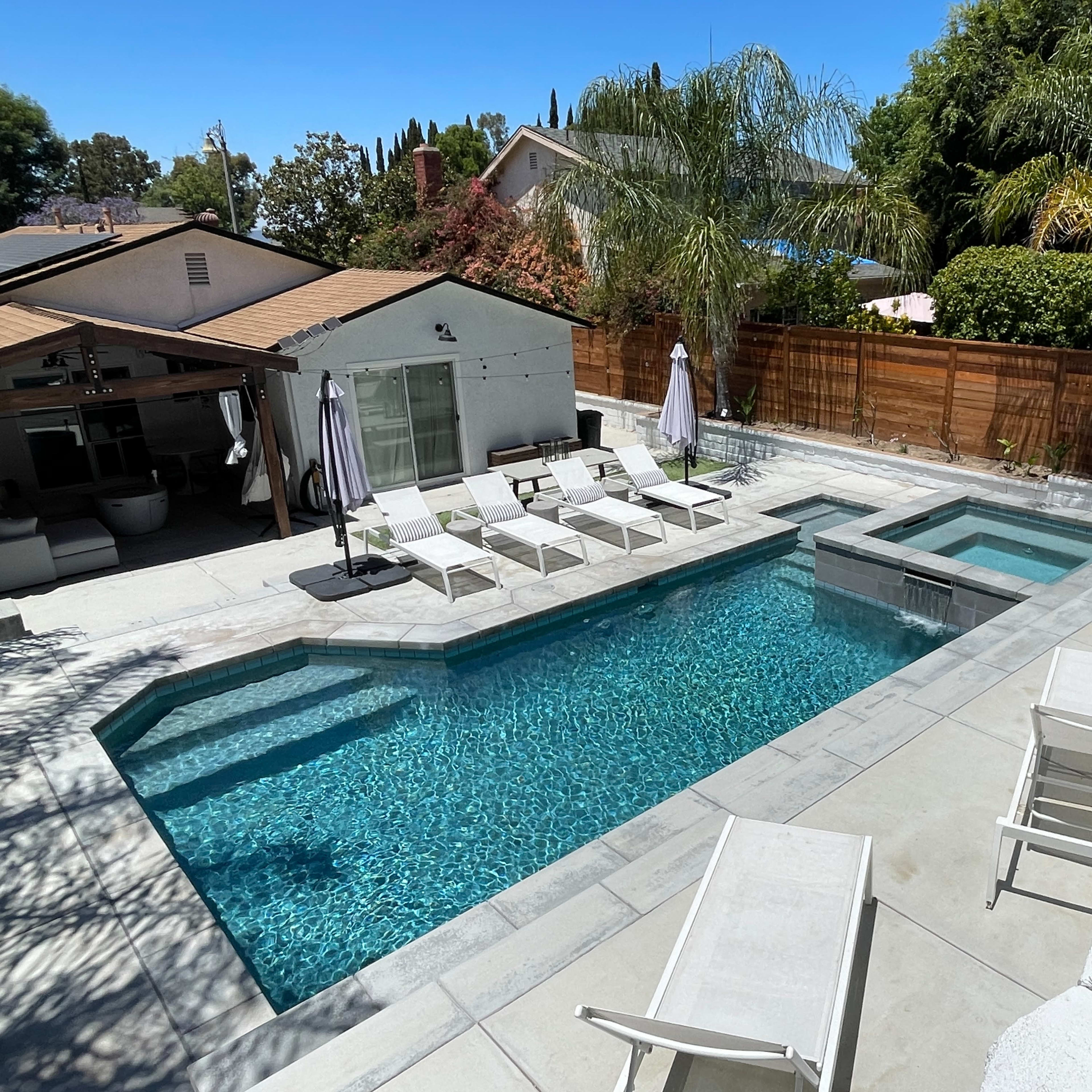 A backyard pool area featuring a rectangular swimming pool with a spa, surrounded by lounge chairs and palm trees.
