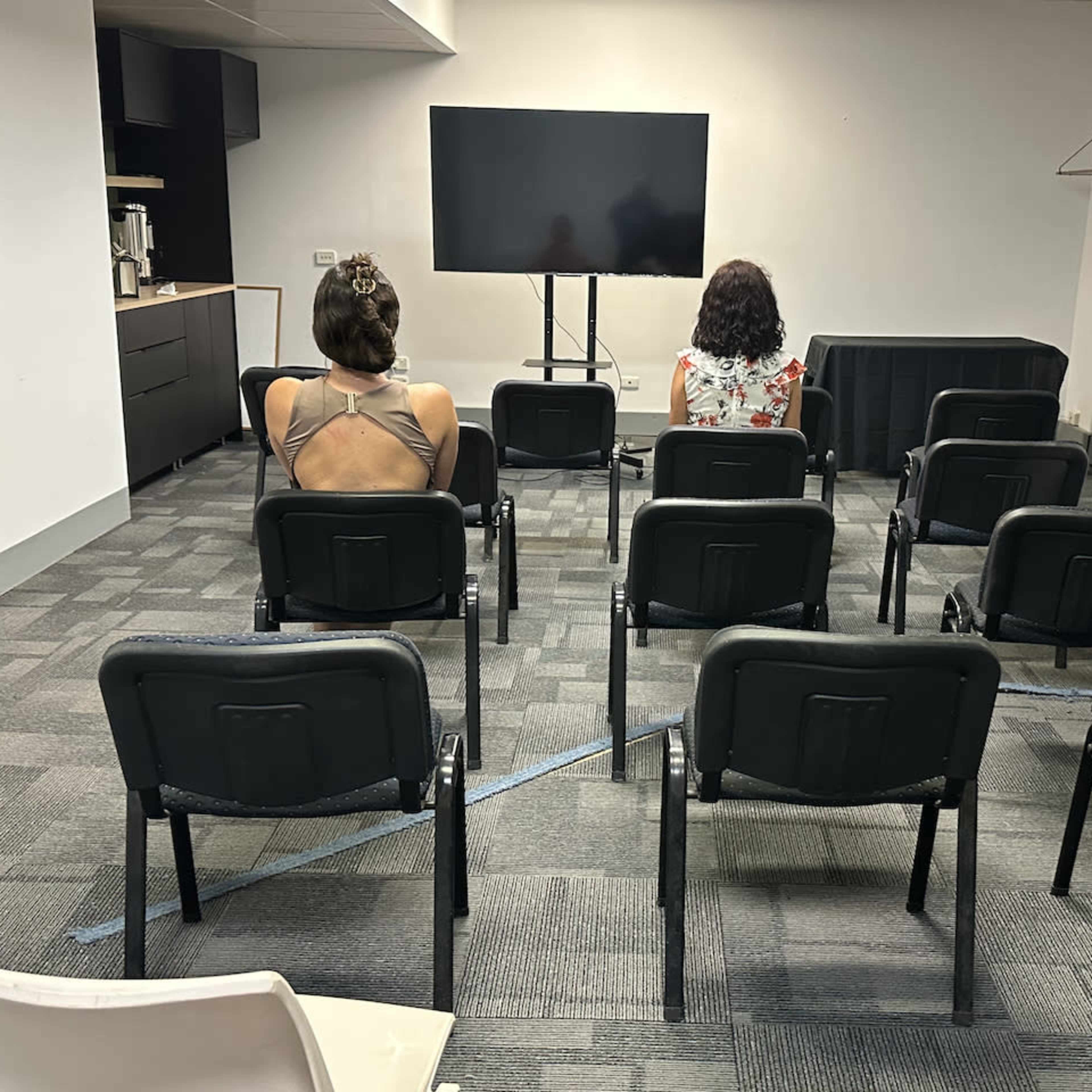 Two individuals sit facing a blank television screen in a sparsely arranged seating area with empty chairs.