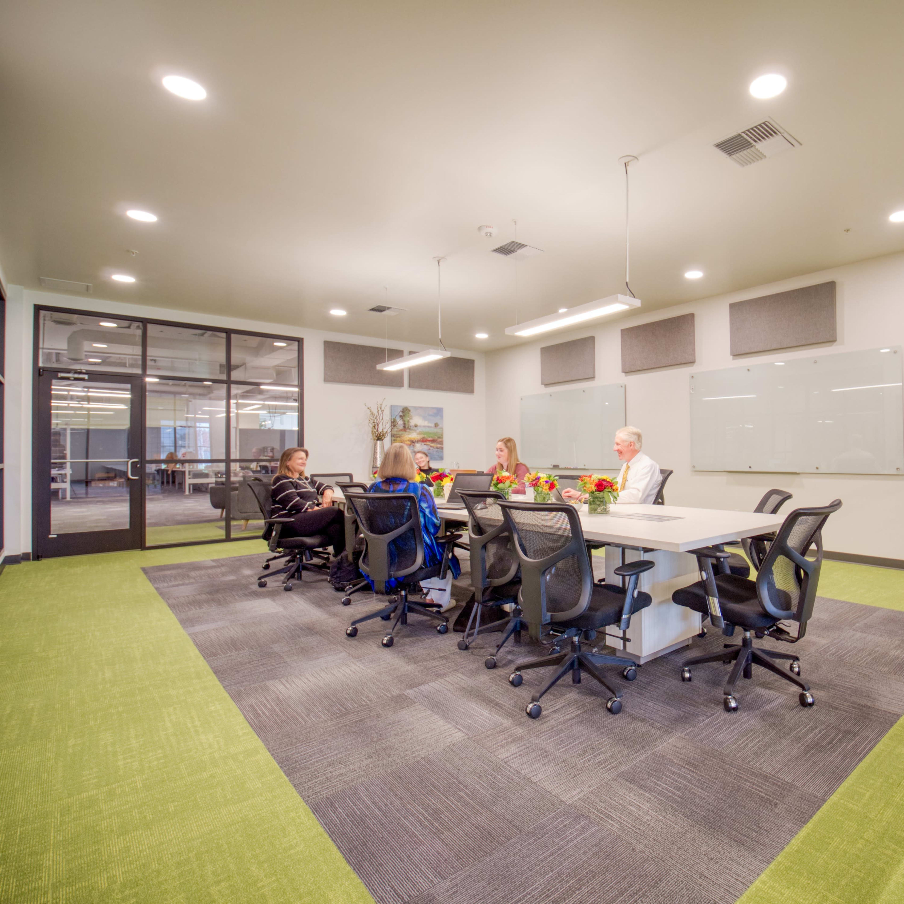 A group of five people sits around a rectangular conference table in an office meeting room with green carpet and white walls.