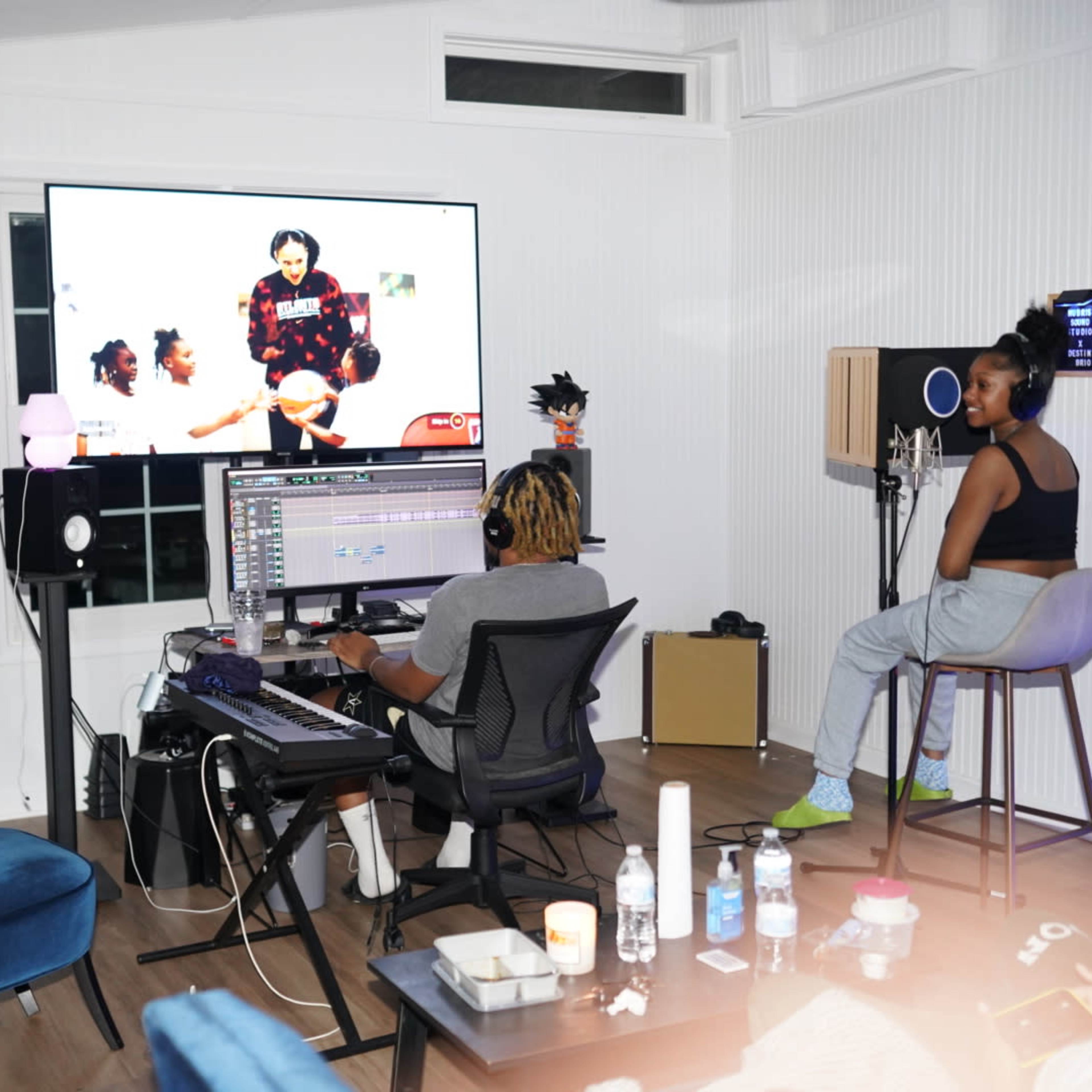 Two individuals are seated in a music studio, one working on a computer with music software, while the other listens through headphones, with a large screen displaying a video.