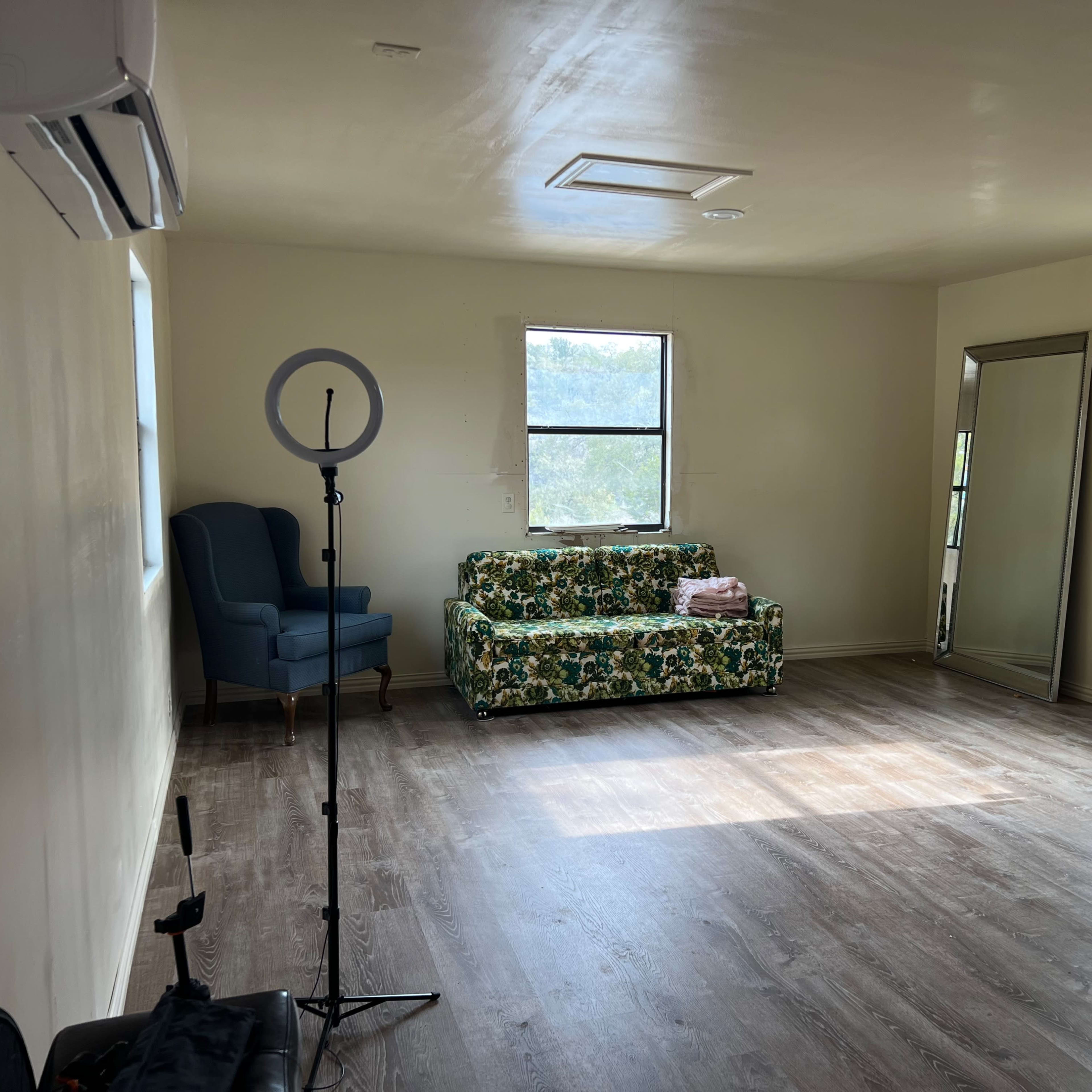 A vacant room with a floral-patterned sofa, a tall mirror, and a ring light setup beside an armchair.