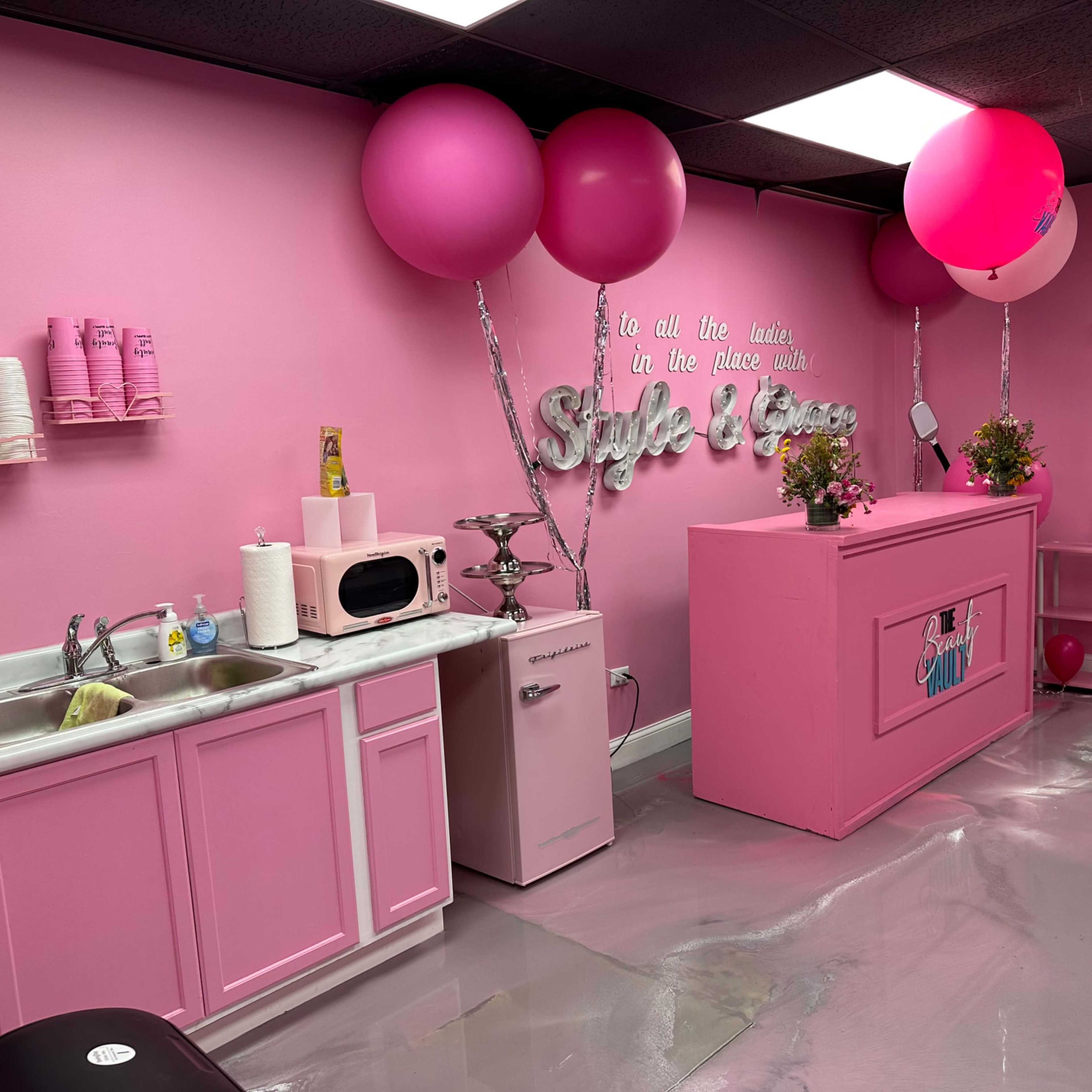 A brightly colored pink interior of a beauty salon with a kitchenette area, decorated with balloons and flowers.