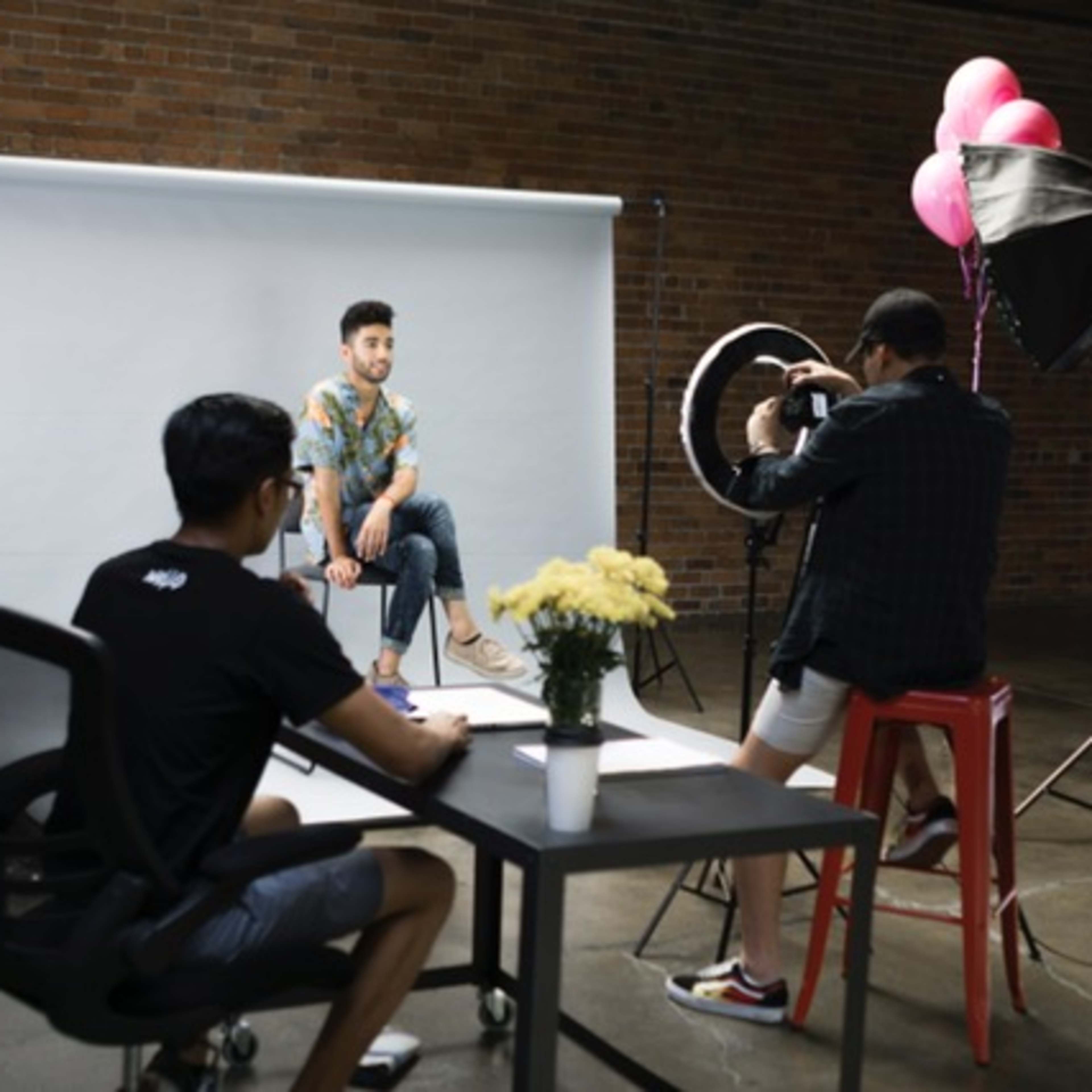 A young man poses for a photoshoot while a photographer captures the scene and another person sits at a desk nearby.