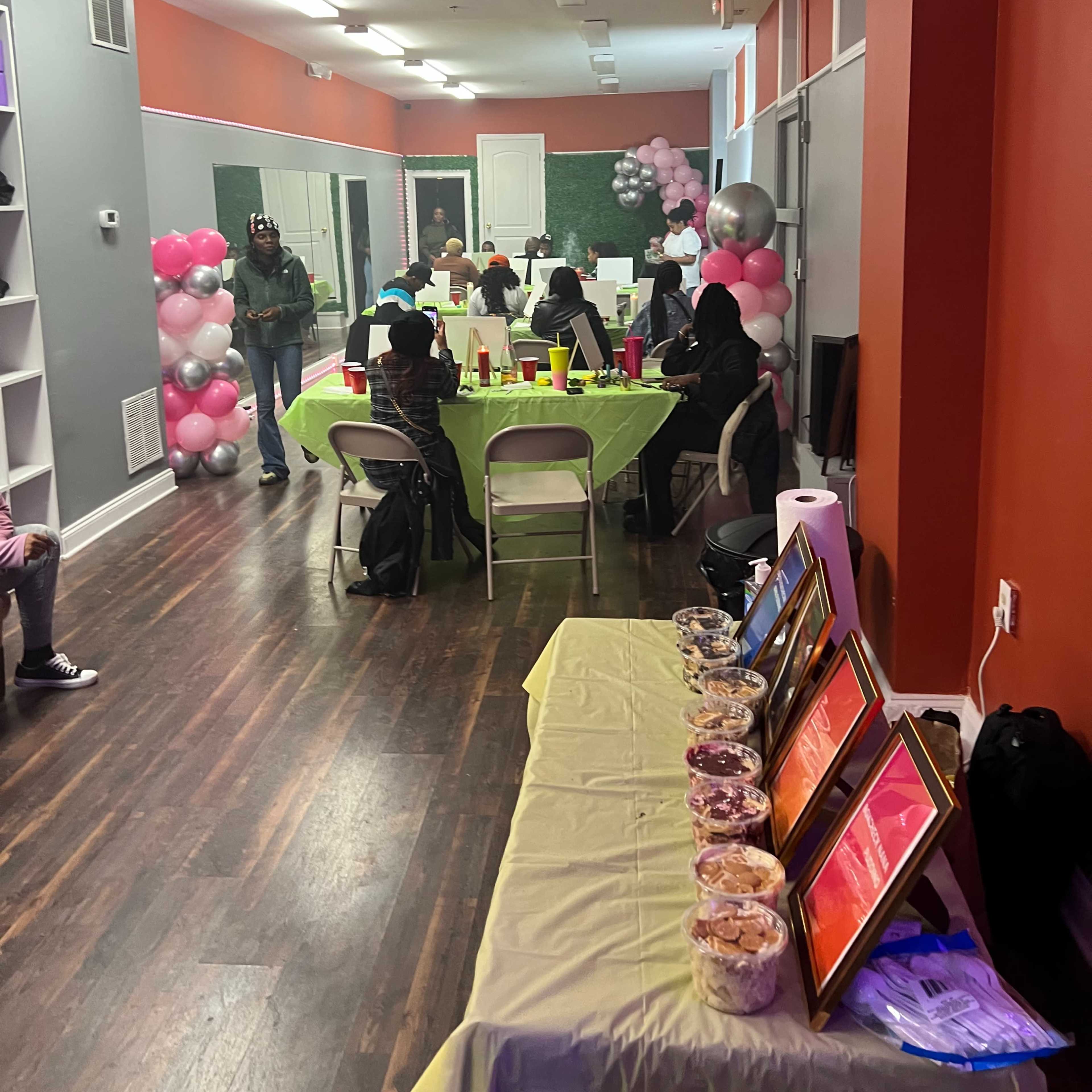 A decorated event space with tables covered in bright green cloth, a buffet of snacks, and balloons along the walls, while guests sit around enjoying the gathering.