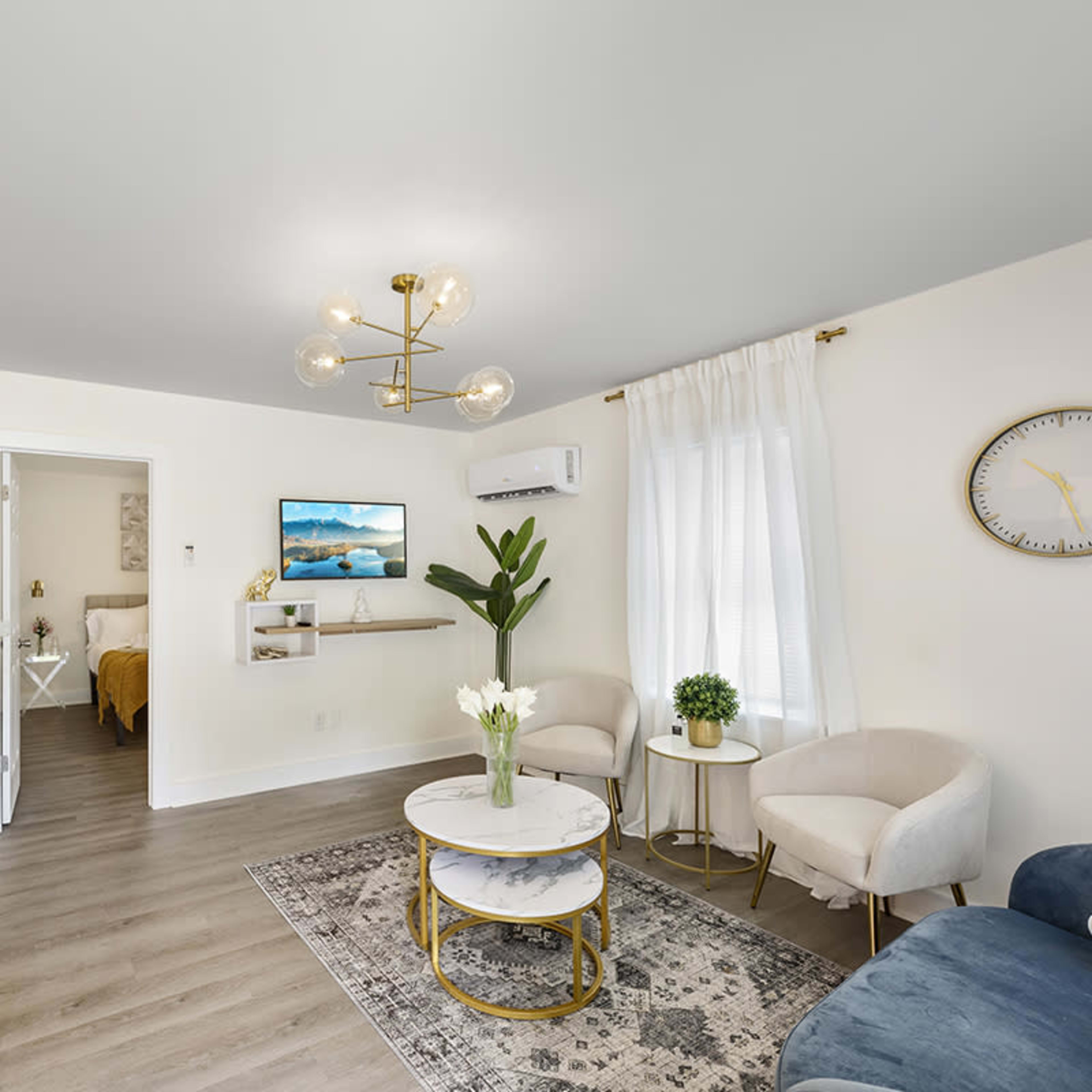 A modern living room with a coffee table, two accent chairs, a wall-mounted television, and a view of a separate bedroom through an open doorway.