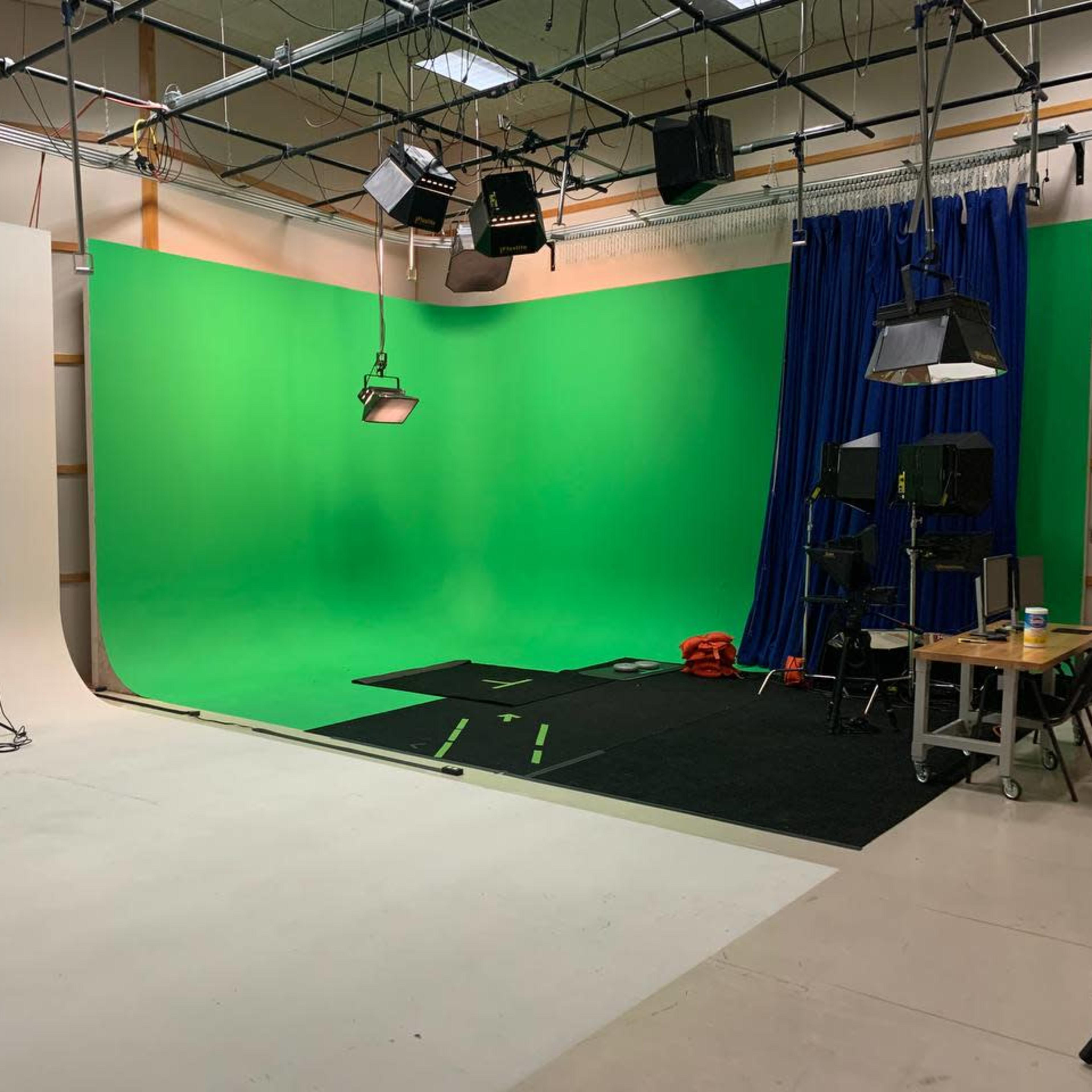 A spacious studio setup featuring a green screen, black carpet, various lighting equipment, and a table with additional items on one side.