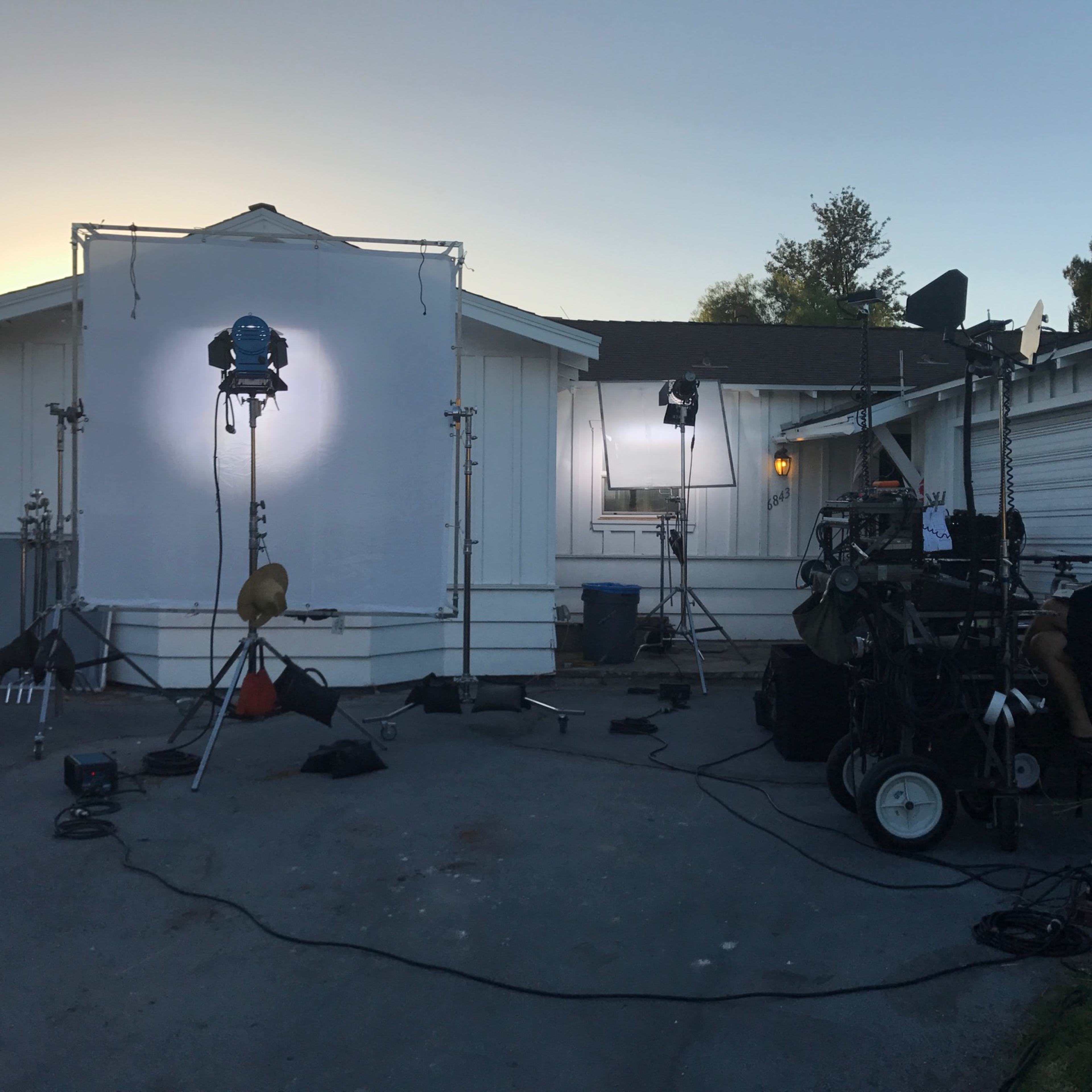 A film set outside a white building, equipped with lights, reflectors, and camera equipment.
