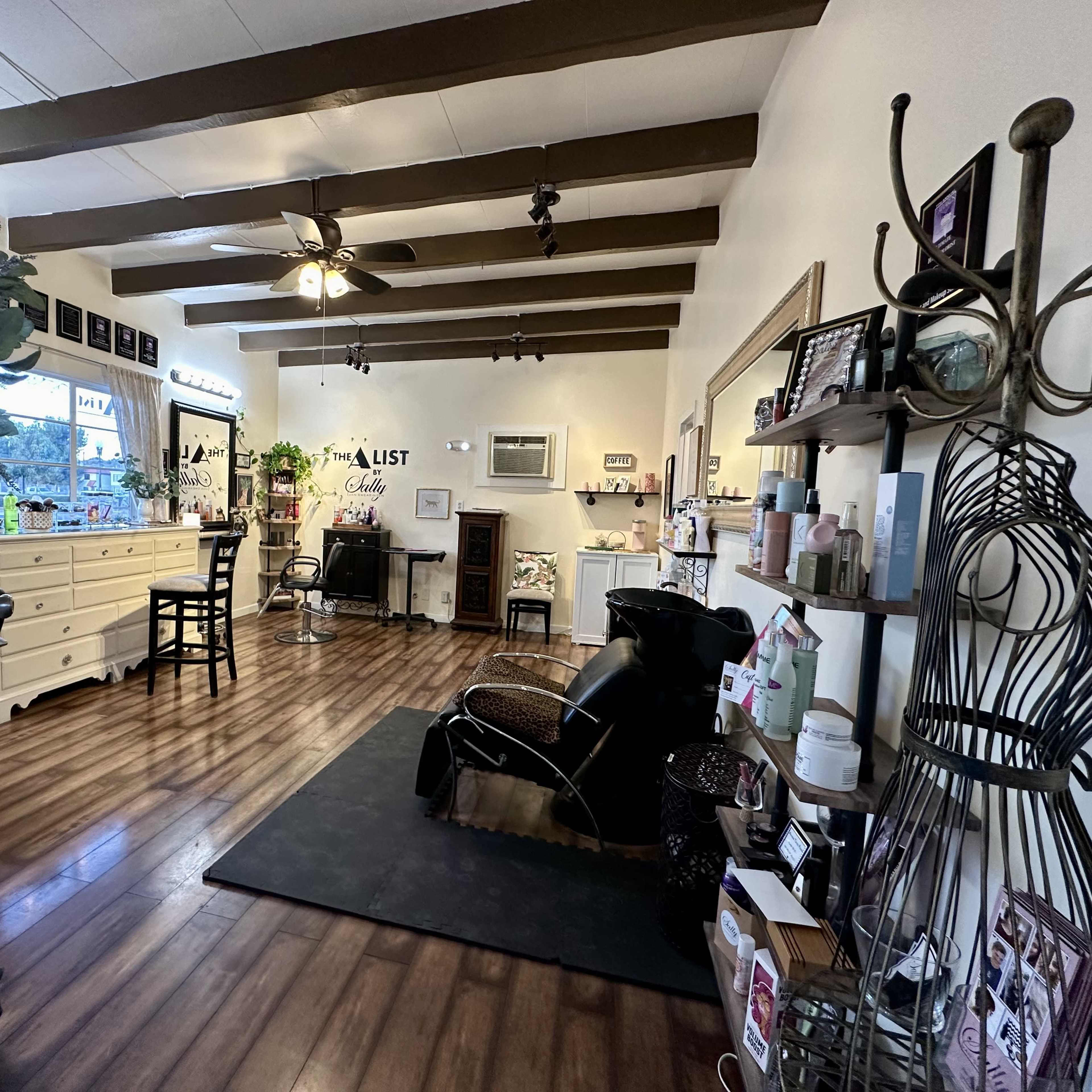A hair salon with wooden beams on the ceiling, a waiting area with a black chair, and various styling stations arranged along the walls.