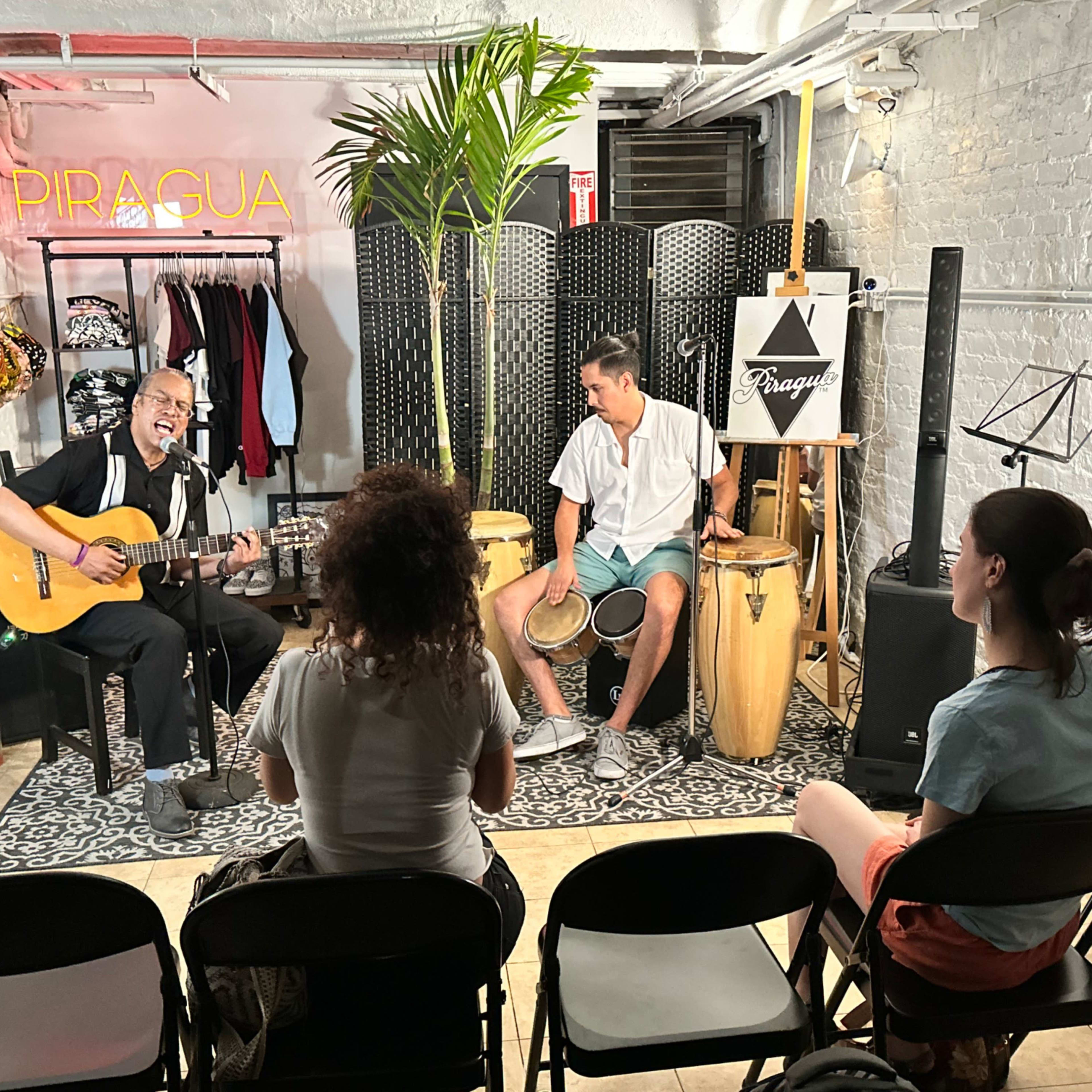 A musician plays guitar while another performs on conga drums in a small indoor venue with chairs facing the stage.