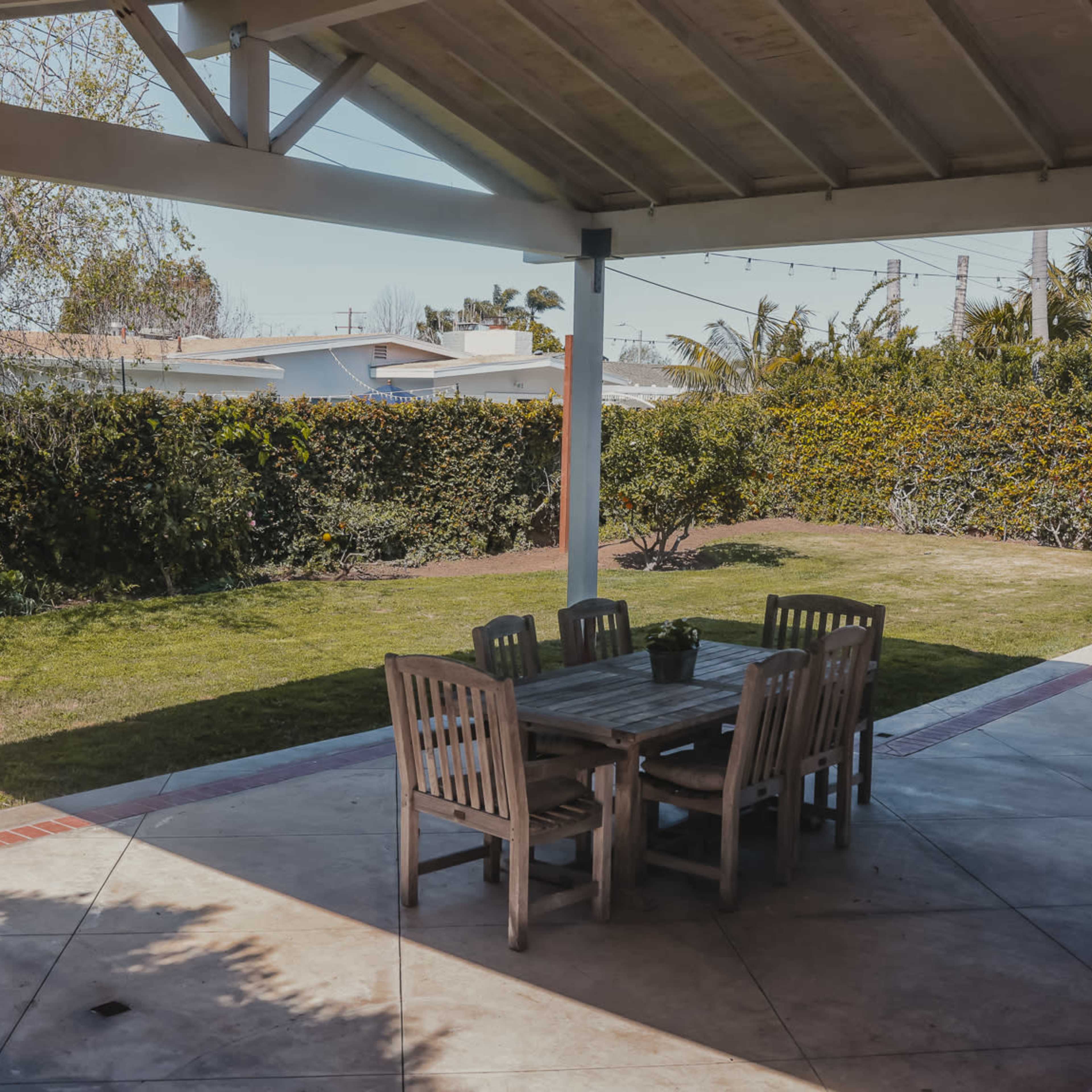 A wooden dining table and chairs are set up under a covered patio overlooking a grassy backyard with hedges and trees.