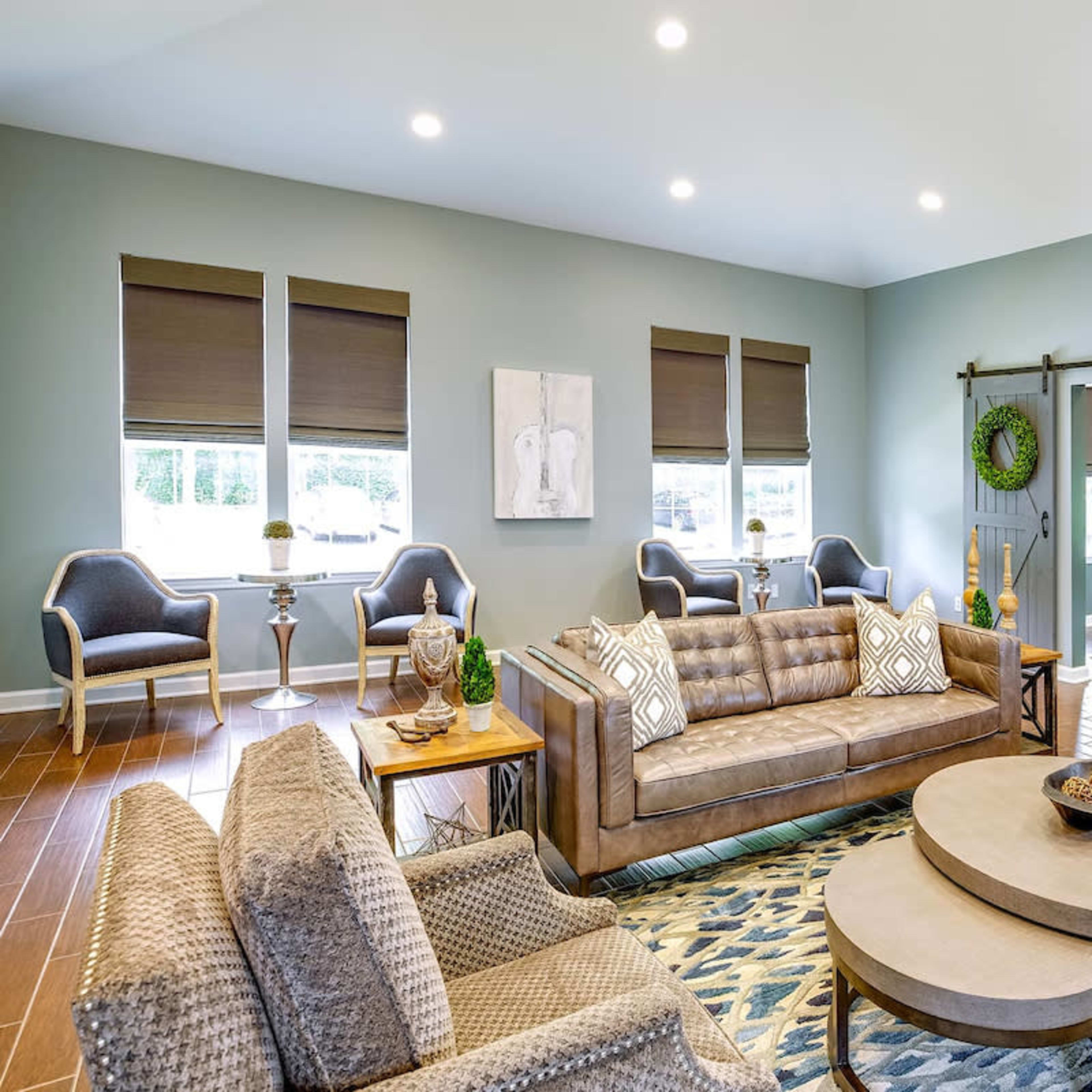A modern living room features a brown sofa and chairs arranged around a coffee table, with windows adorned by dark shades and decorative wall accents.