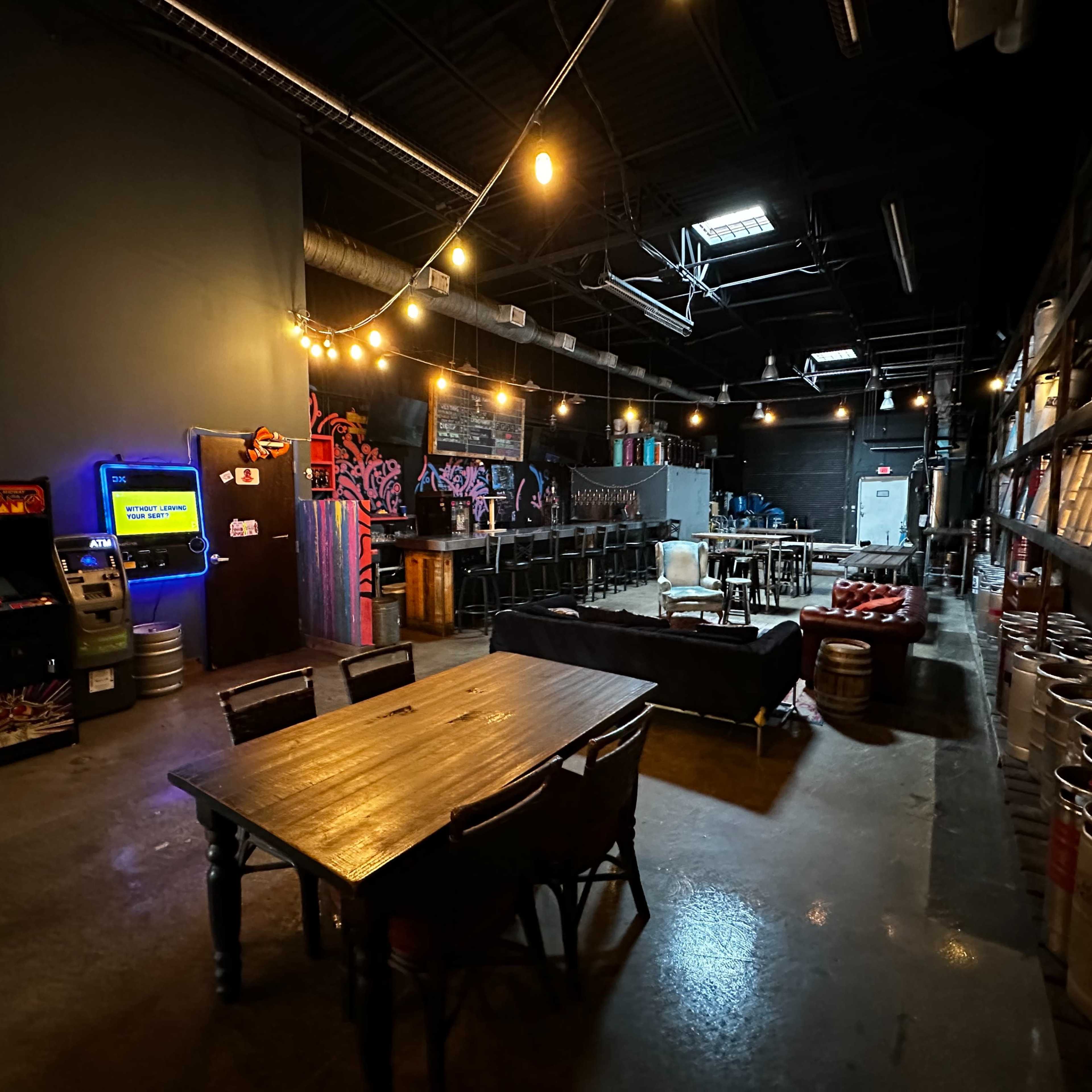 A spacious, dimly lit bar area with a long table, arcade games, a counter with bar stools, and shelves lined with kegs.