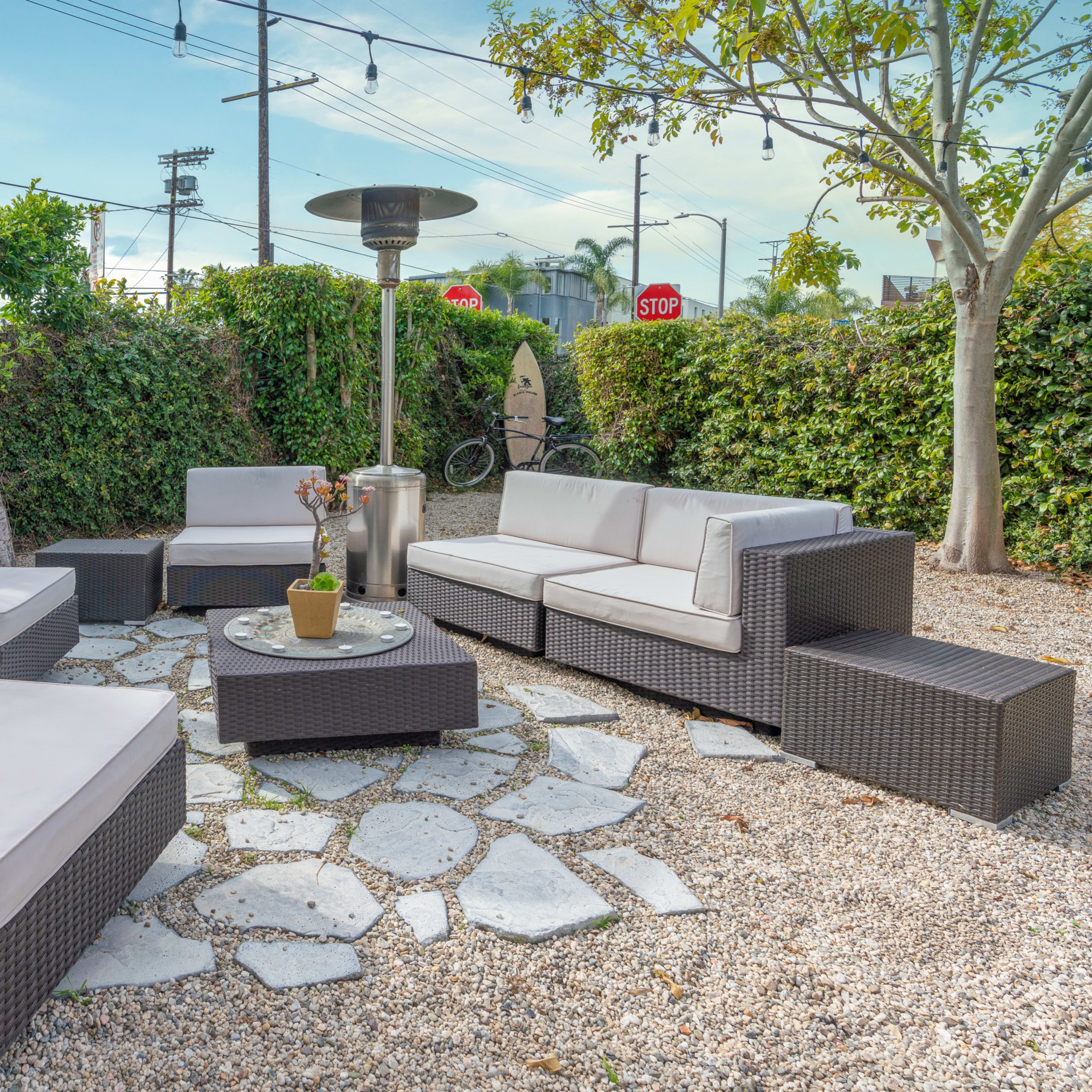 A patio area with modern wicker furniture arranged around a stone table, surrounded by greenery and patio heaters.