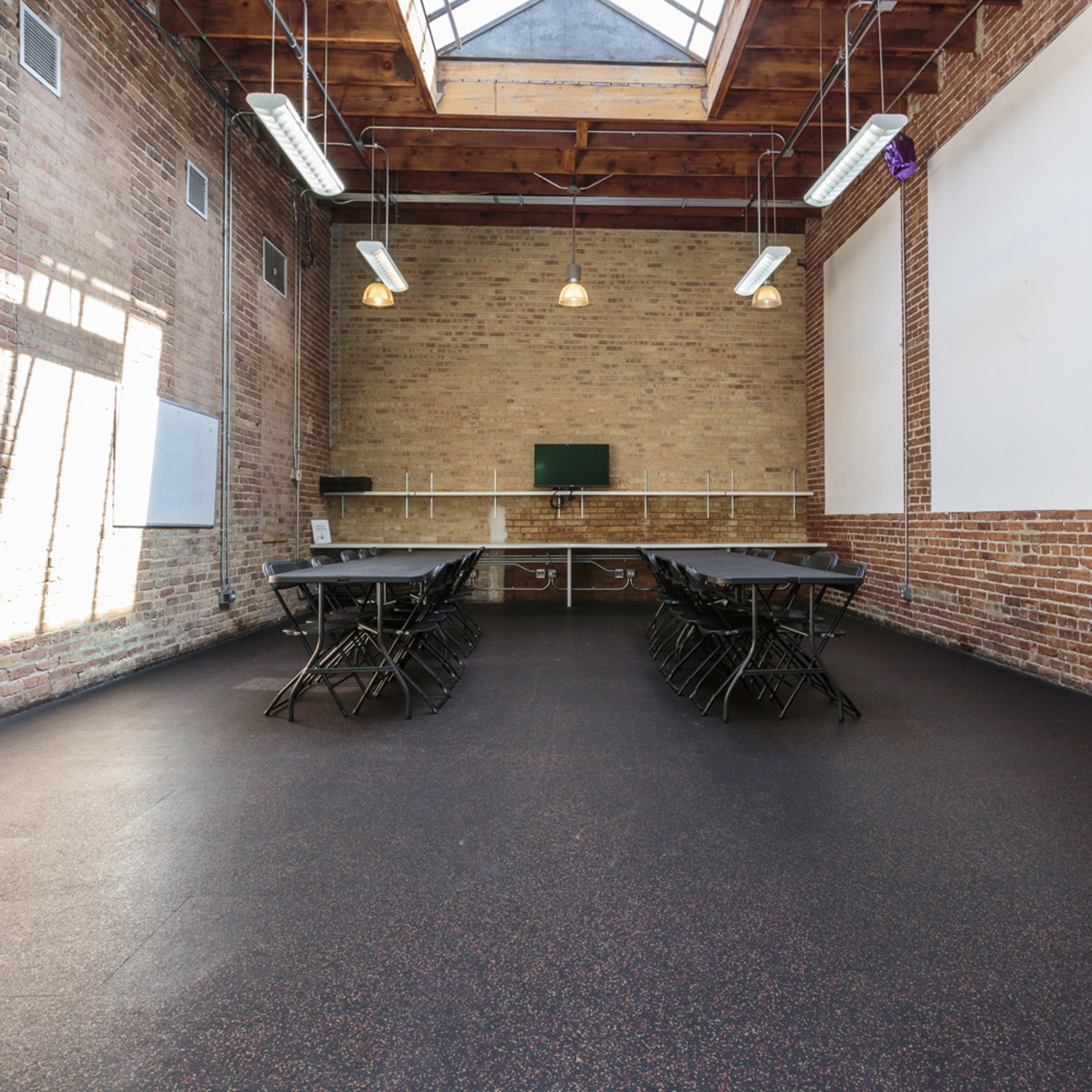 A spacious room with brick walls, a skylight, and several folding tables arranged in rows, along with stacked chairs.