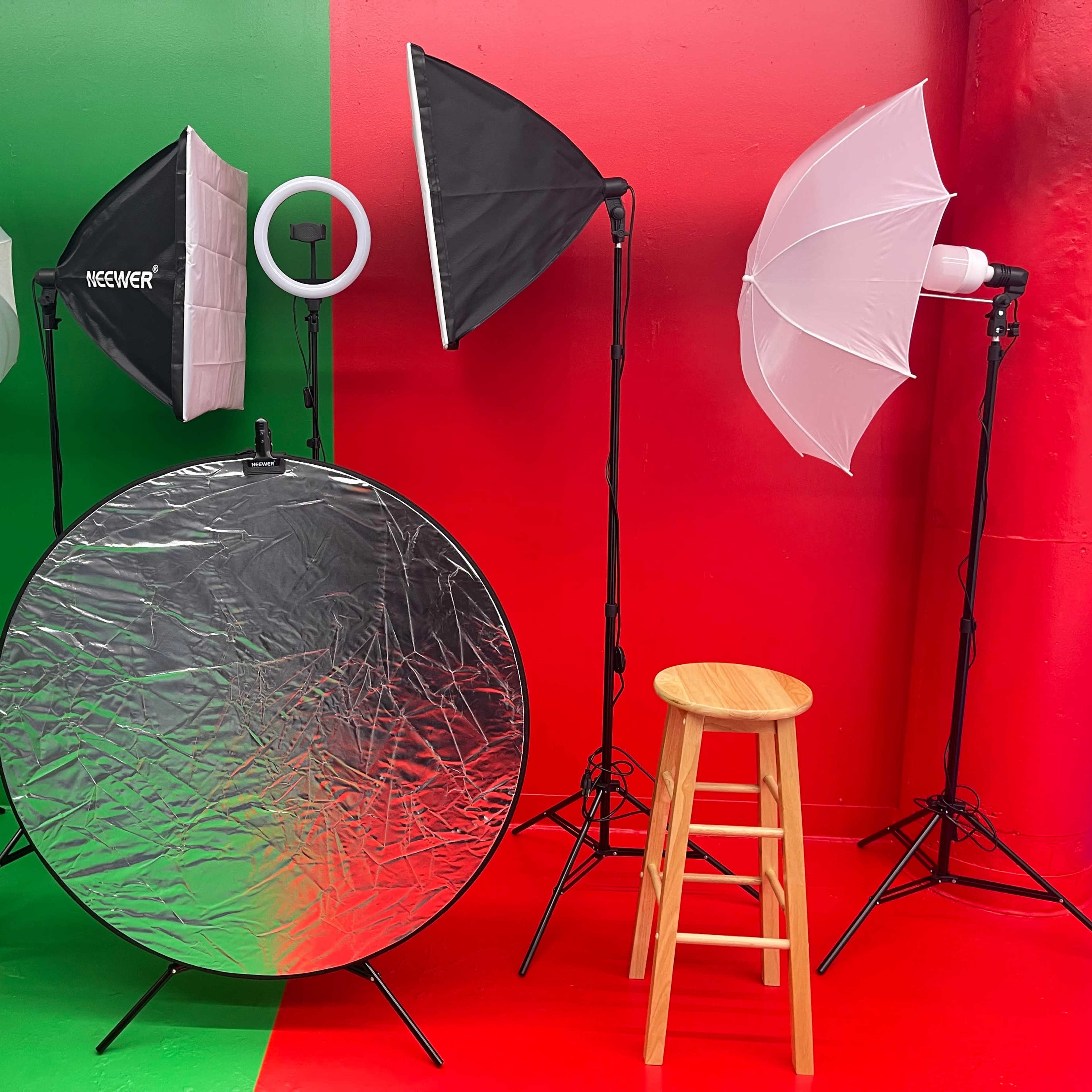 A photography setup with two wooden stools, three light stands with softboxes, a ring light, and a reflective silver disk against a red and green background.