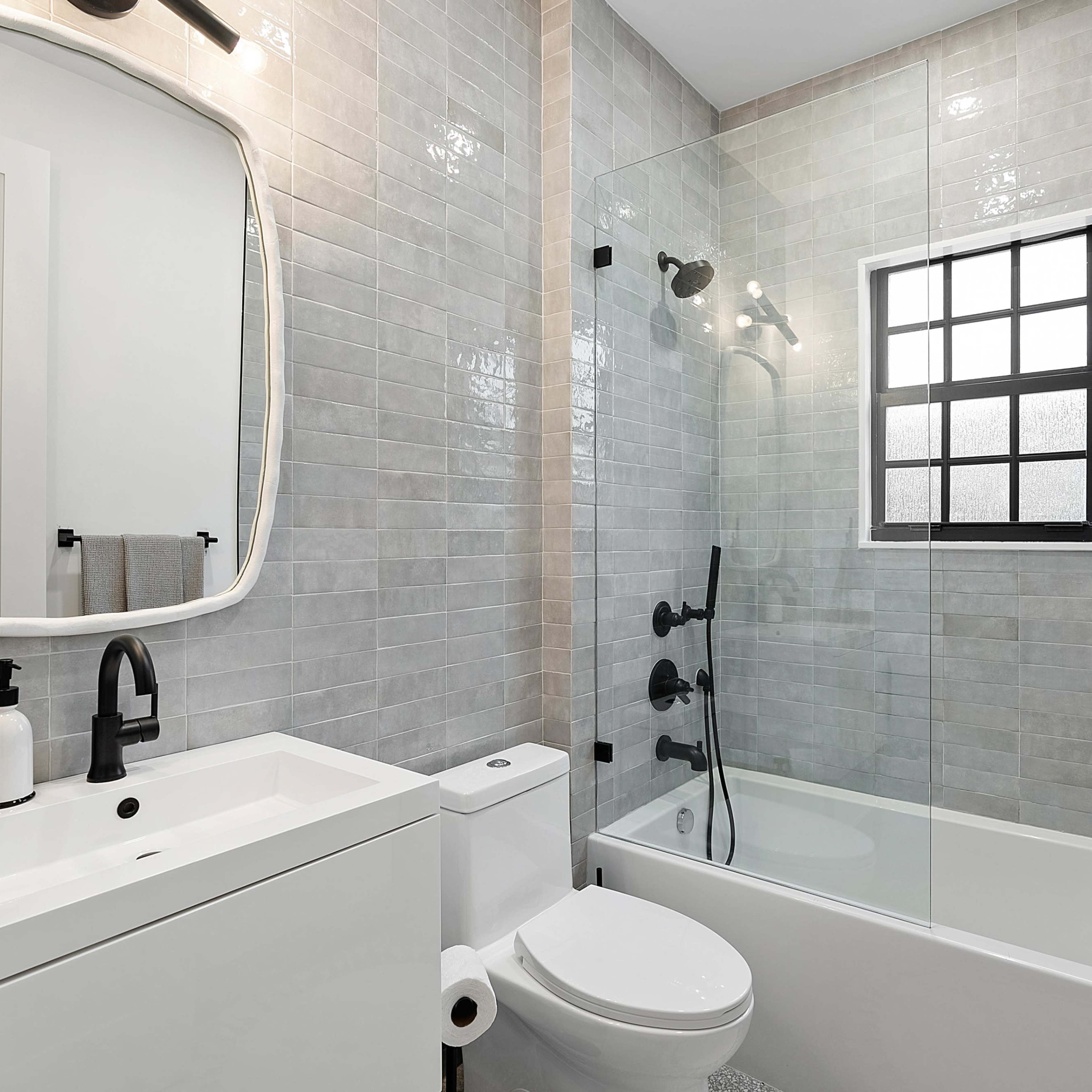 A modern bathroom featuring a bathtub with a glass enclosure, a wall-mounted sink with a round mirror, and gray tiled walls.
