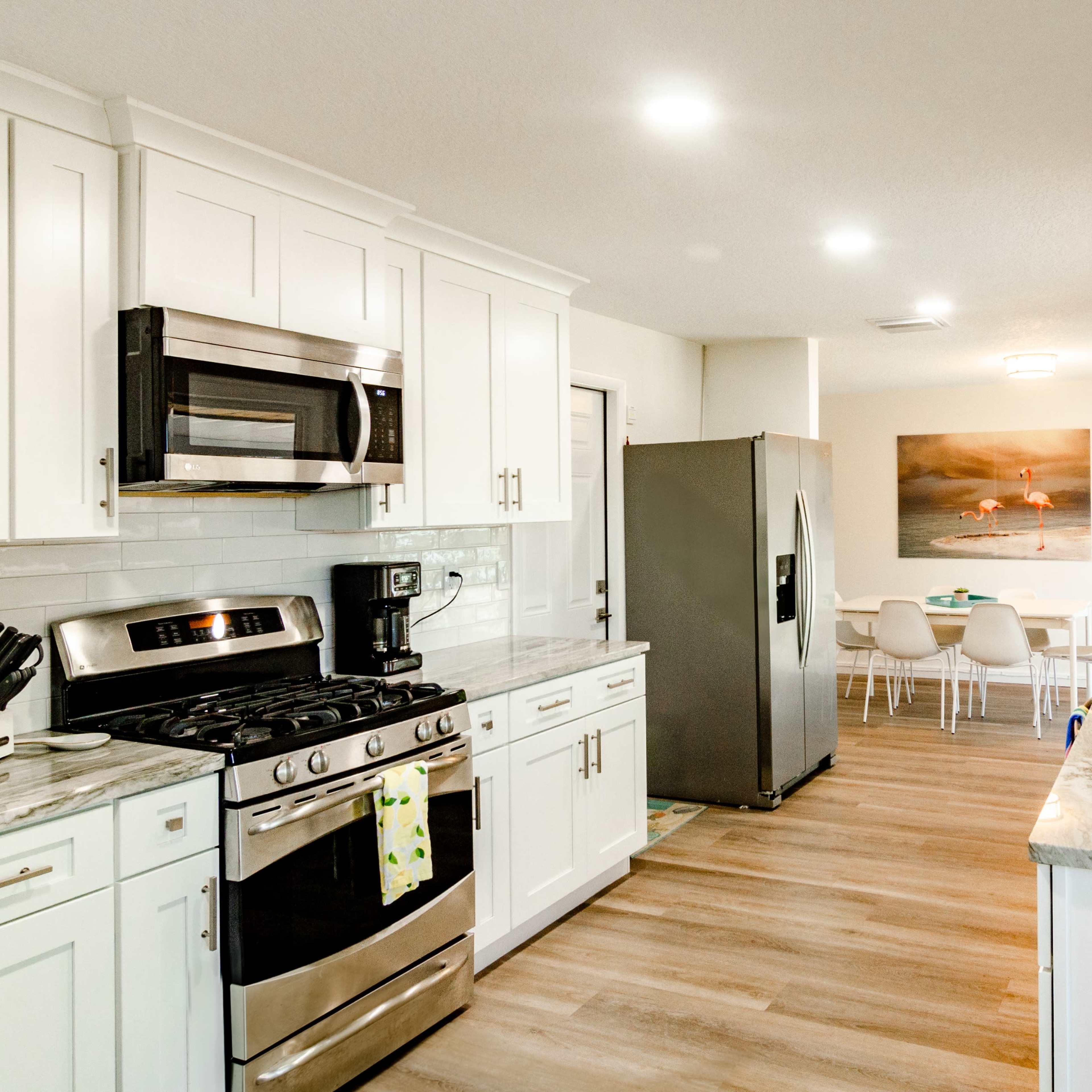 A modern kitchen features white cabinetry, stainless steel appliances, and a marble countertop with a dining area visible in the background.