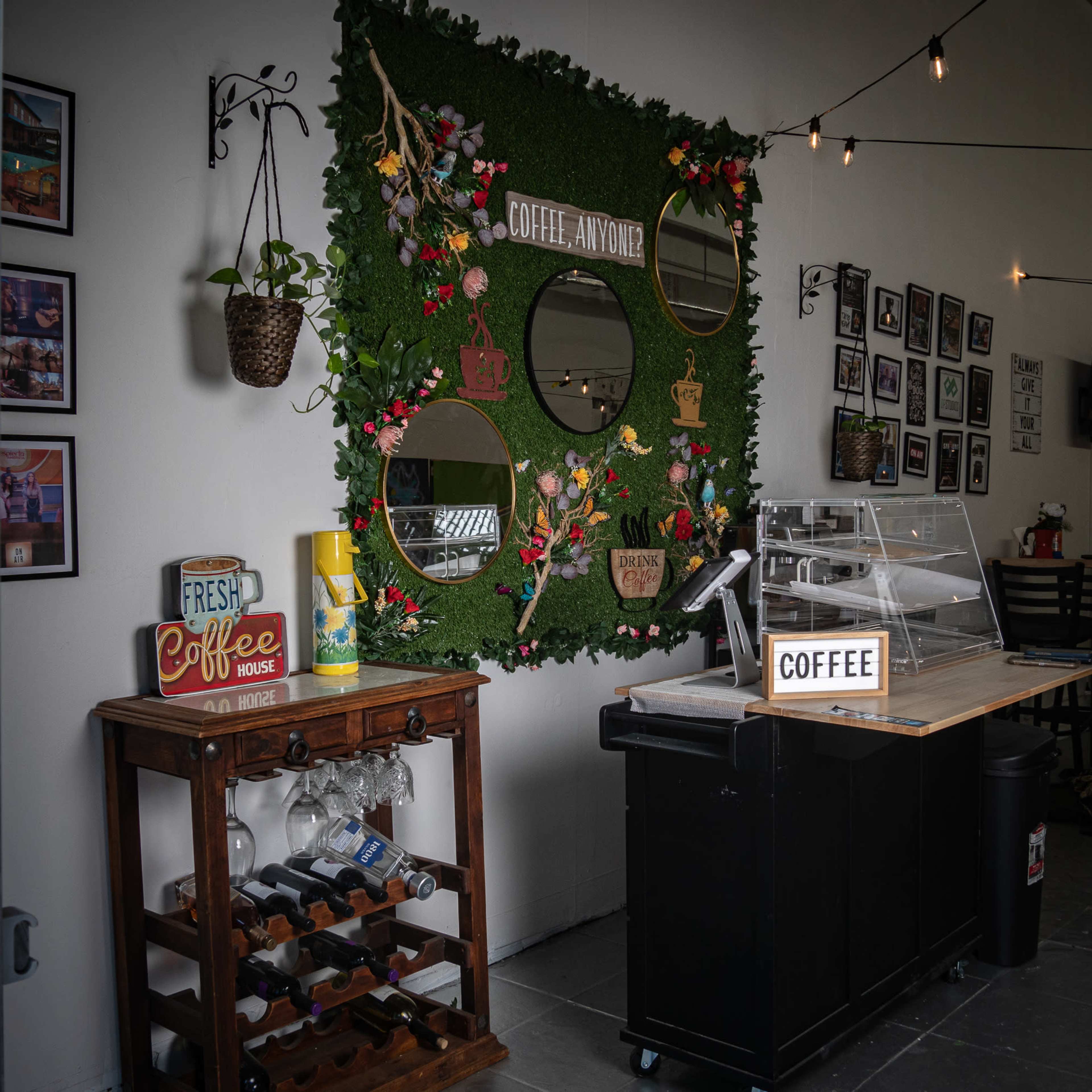 A small café area with a green wall featuring circular mirrors, decorative foliage, and a display counter with a coffee sign and a wine rack.