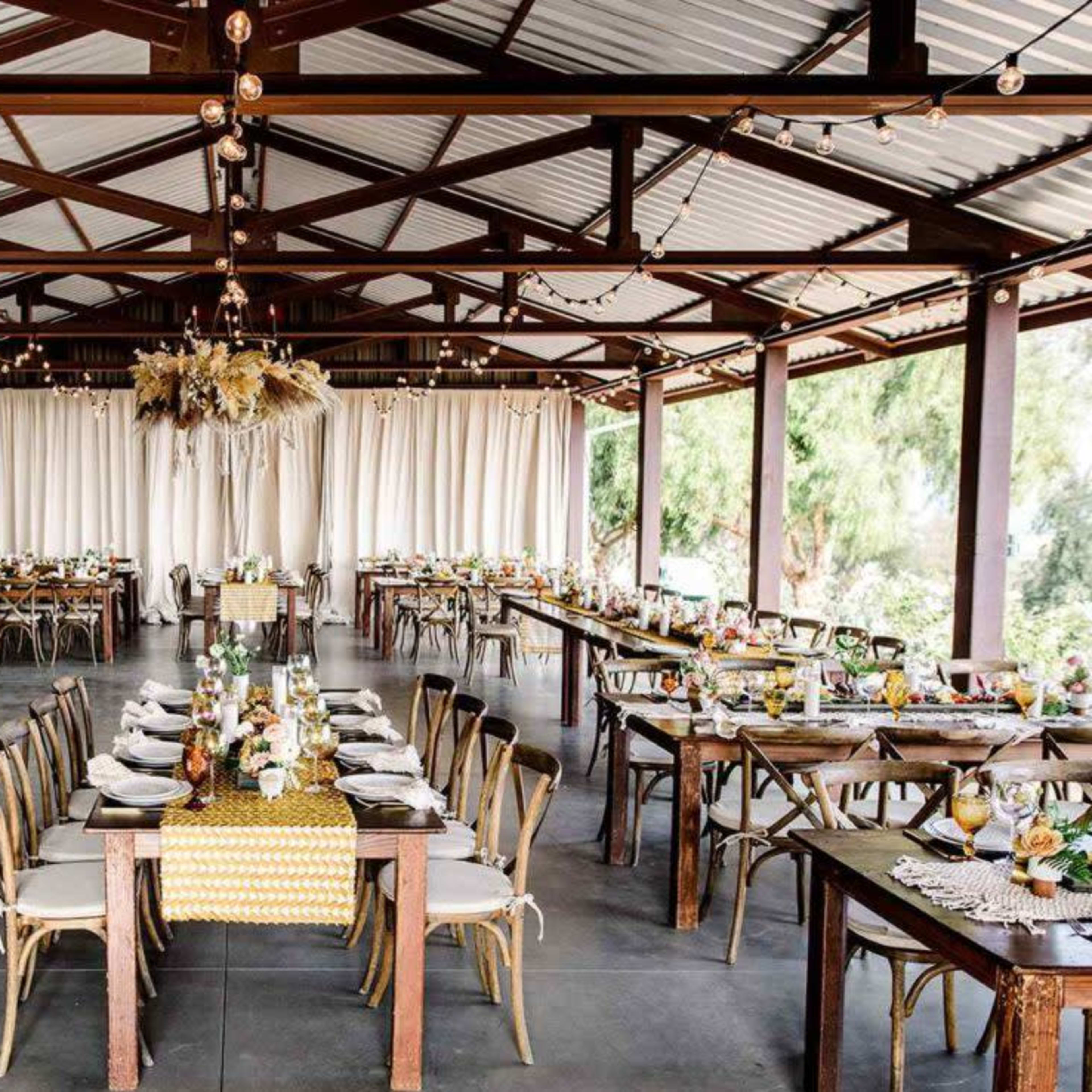 A spacious event venue features rows of wooden tables set with tableware, surrounded by a fabric-draped backdrop and hanging string lights.