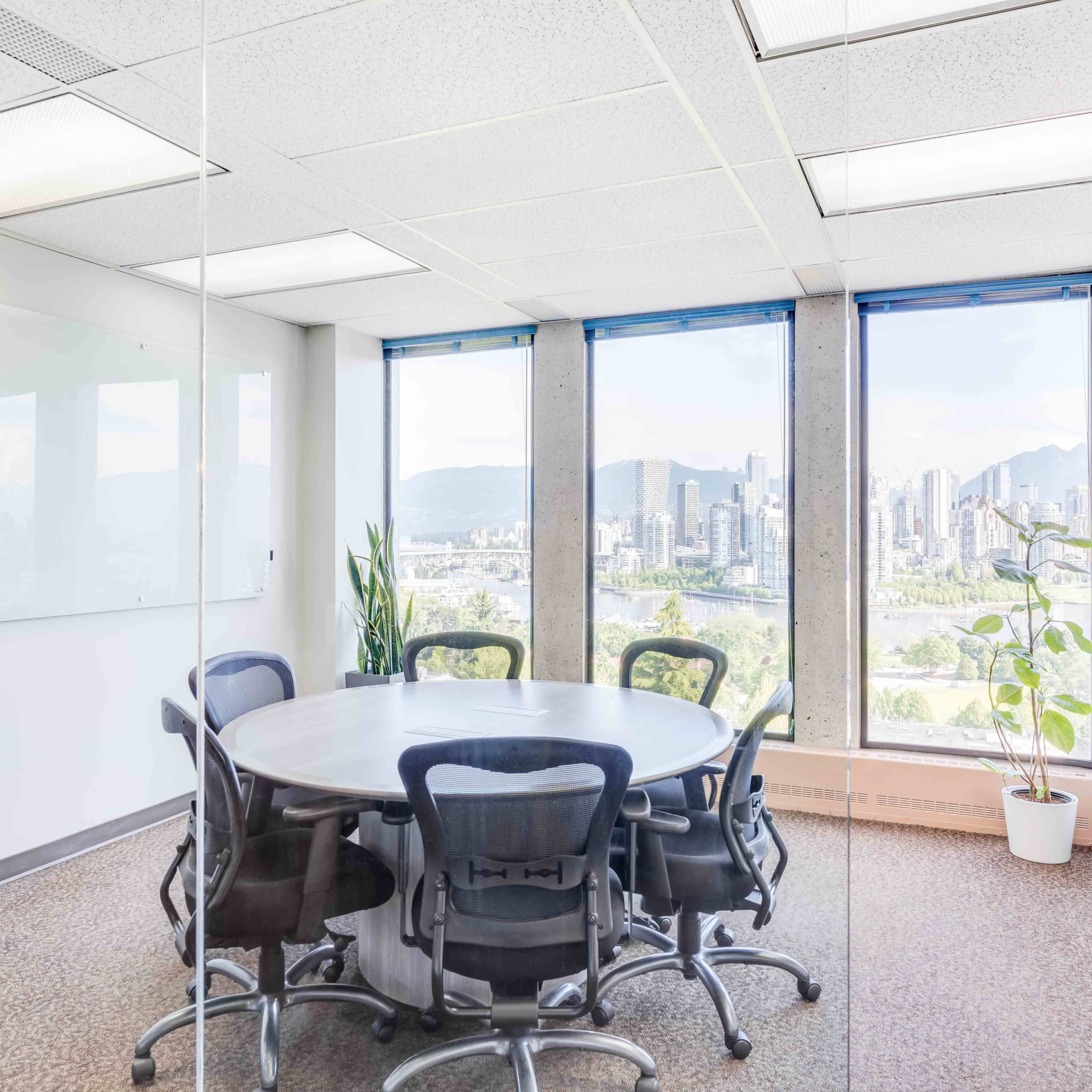A conference room with a round table and chairs overlooks a cityscape through large windows.