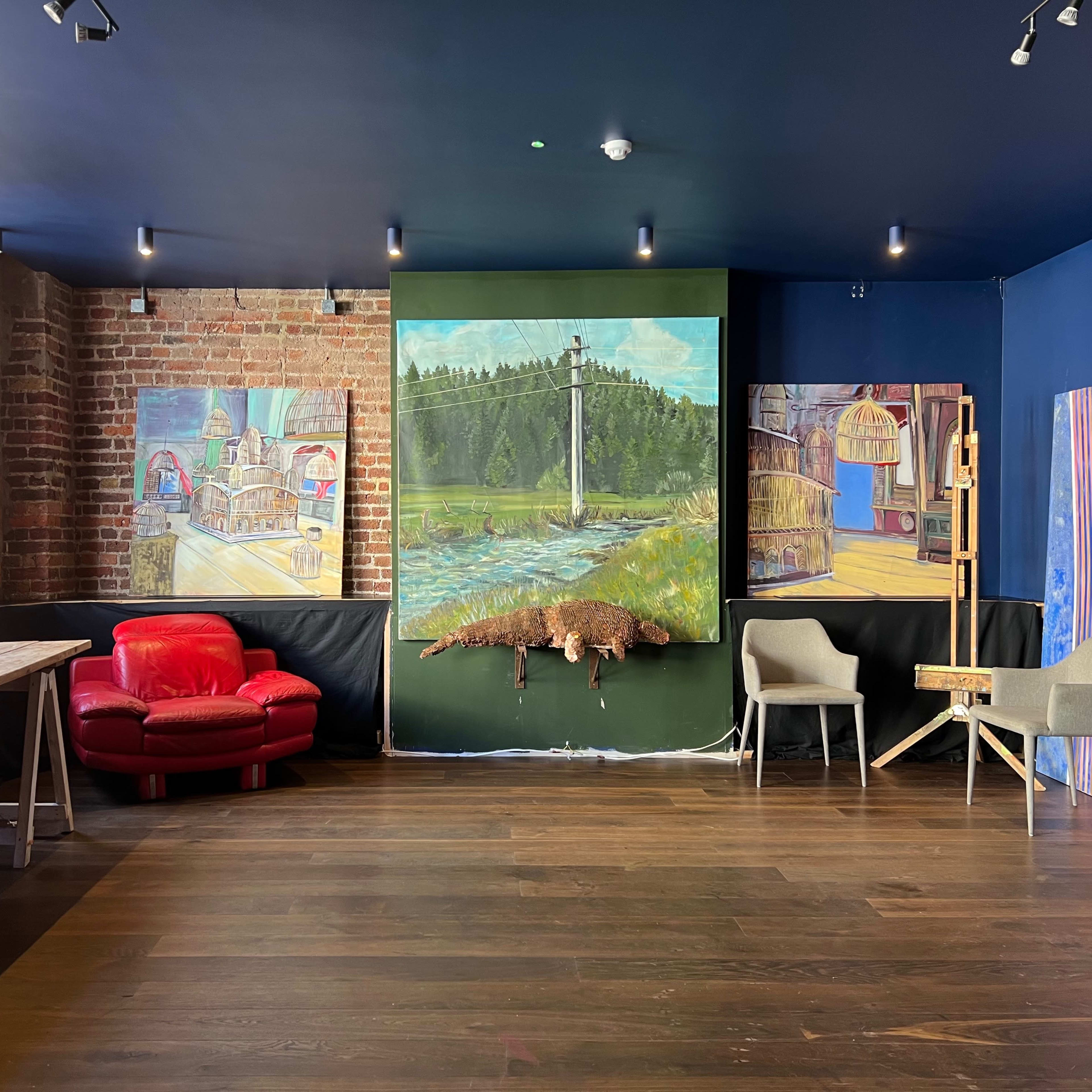 A room with exposed brick walls, featuring a red couch, a wooden table, two chairs, and several large paintings, one depicting a landscape.