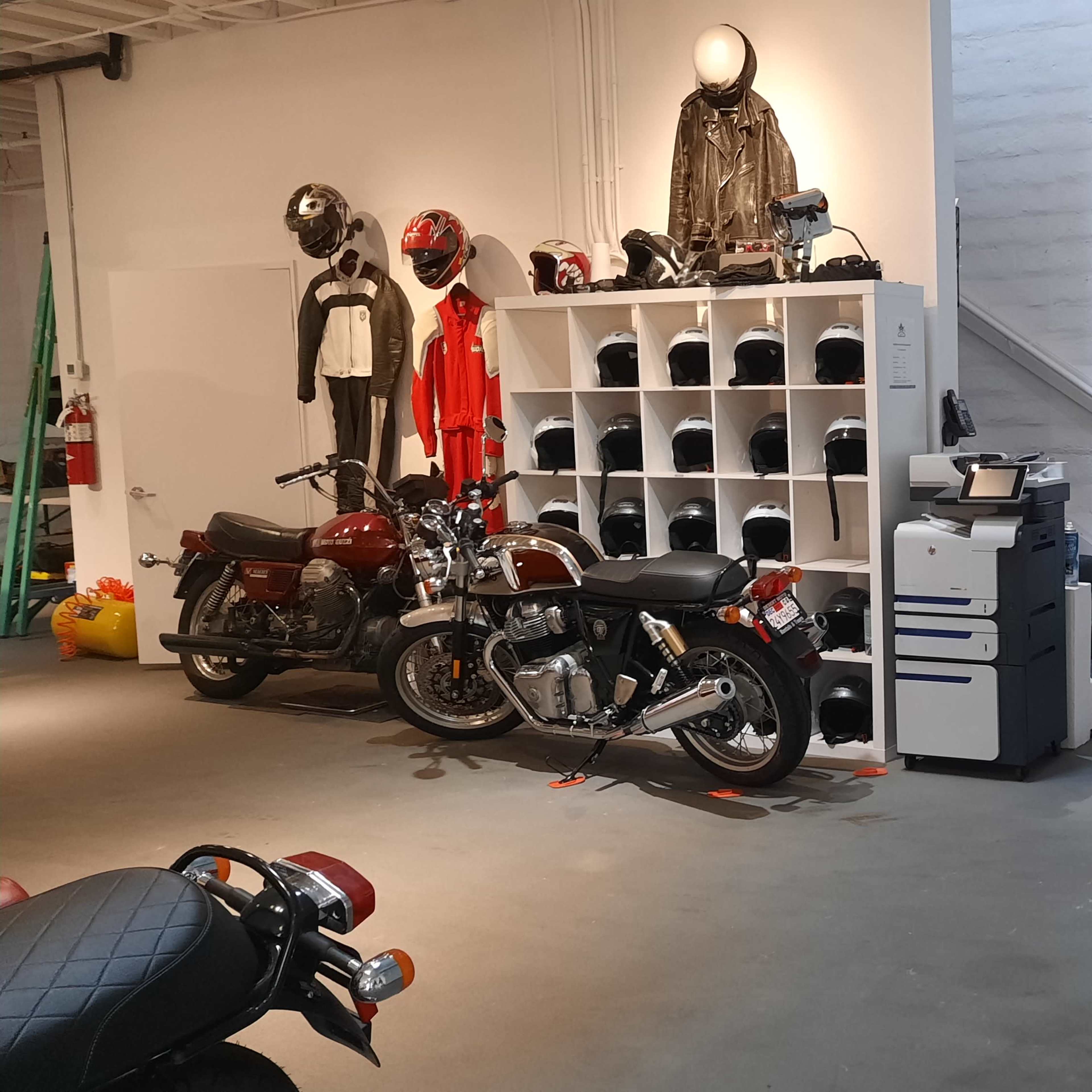 A motorcycle garage featuring two parked motorcycles, a helmet storage unit, a printer, and motorcycle gear hanging on the walls.