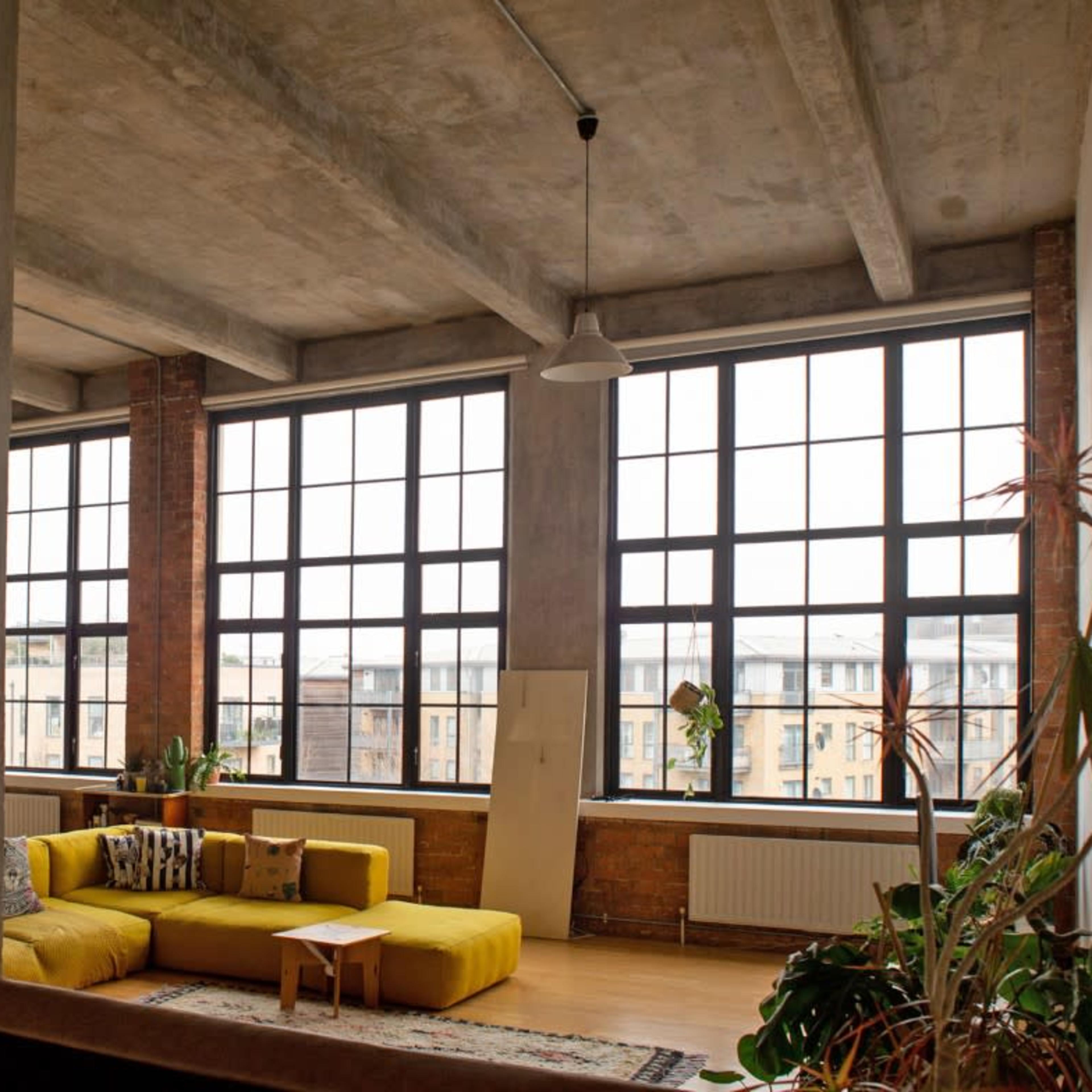 A spacious urban loft with large windows, exposed concrete beams, and casual furniture arranged around a central area.