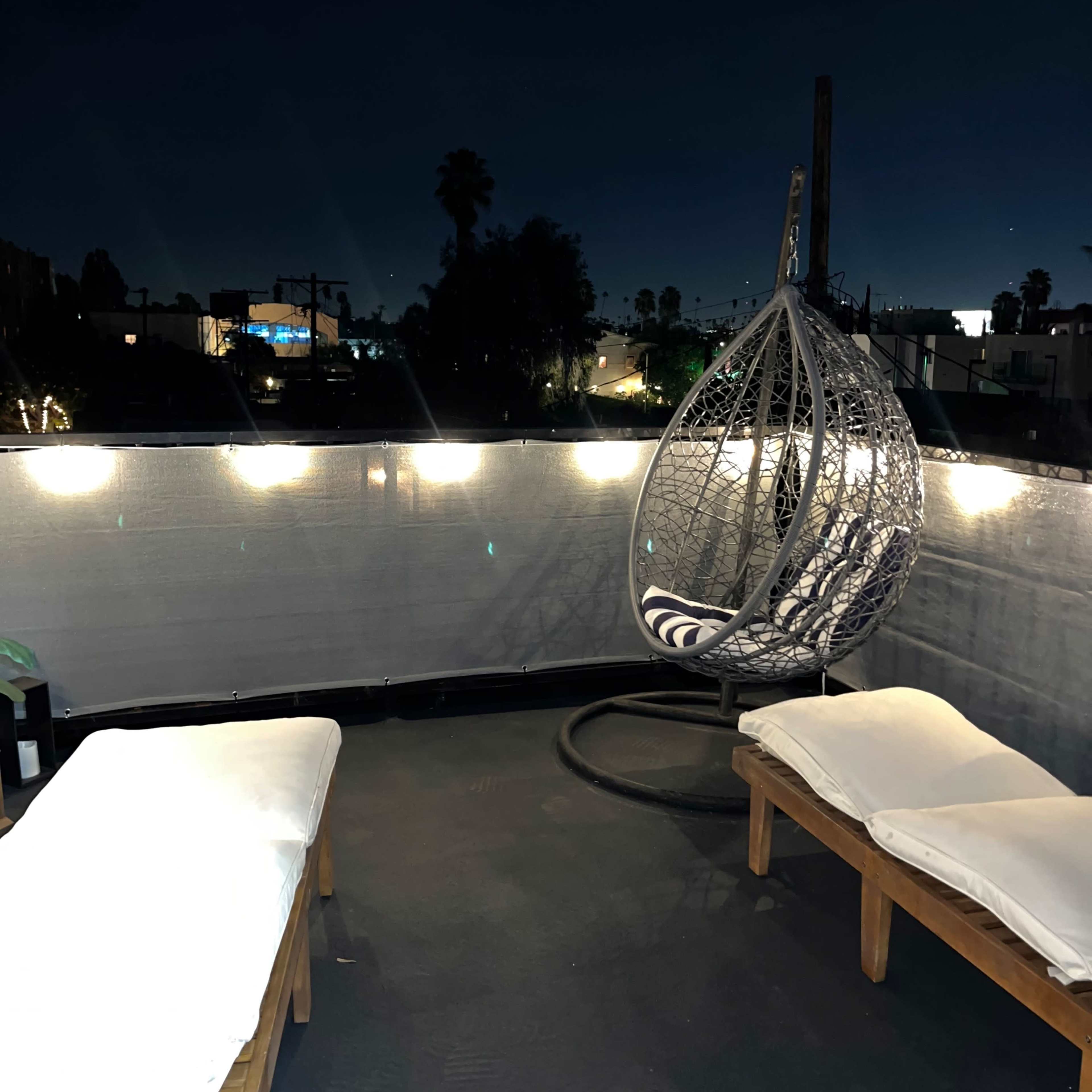 A rooftop terrace features a hanging chair and two loungers, illuminated by string lights against a night sky.