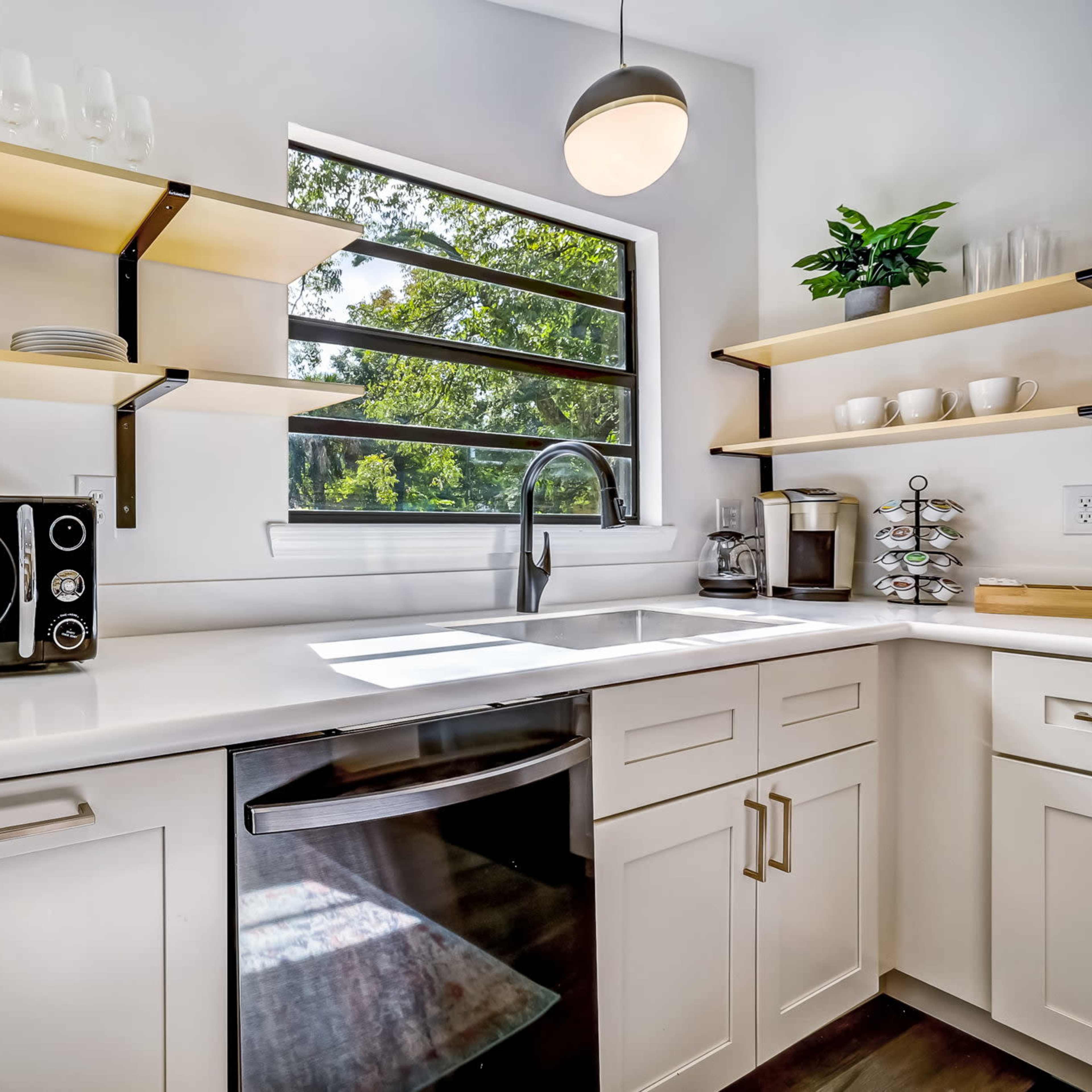 A modern kitchen featuring a stainless steel sink, a microwave, open shelving with plates, and a window allowing natural light to enter.