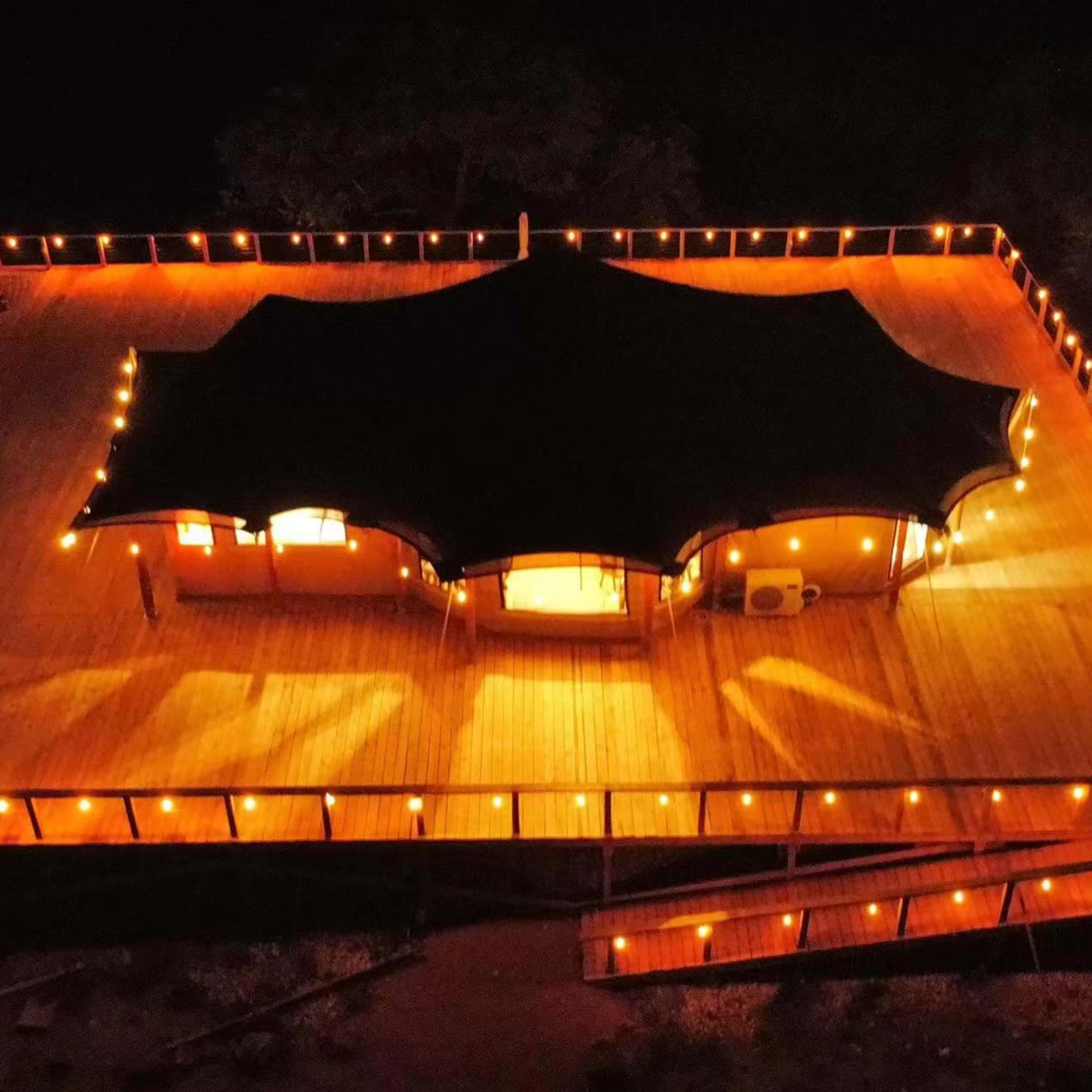 A well-lit, spacious wooden deck surrounds a central tented structure, illuminated by string lights against the nighttime backdrop.