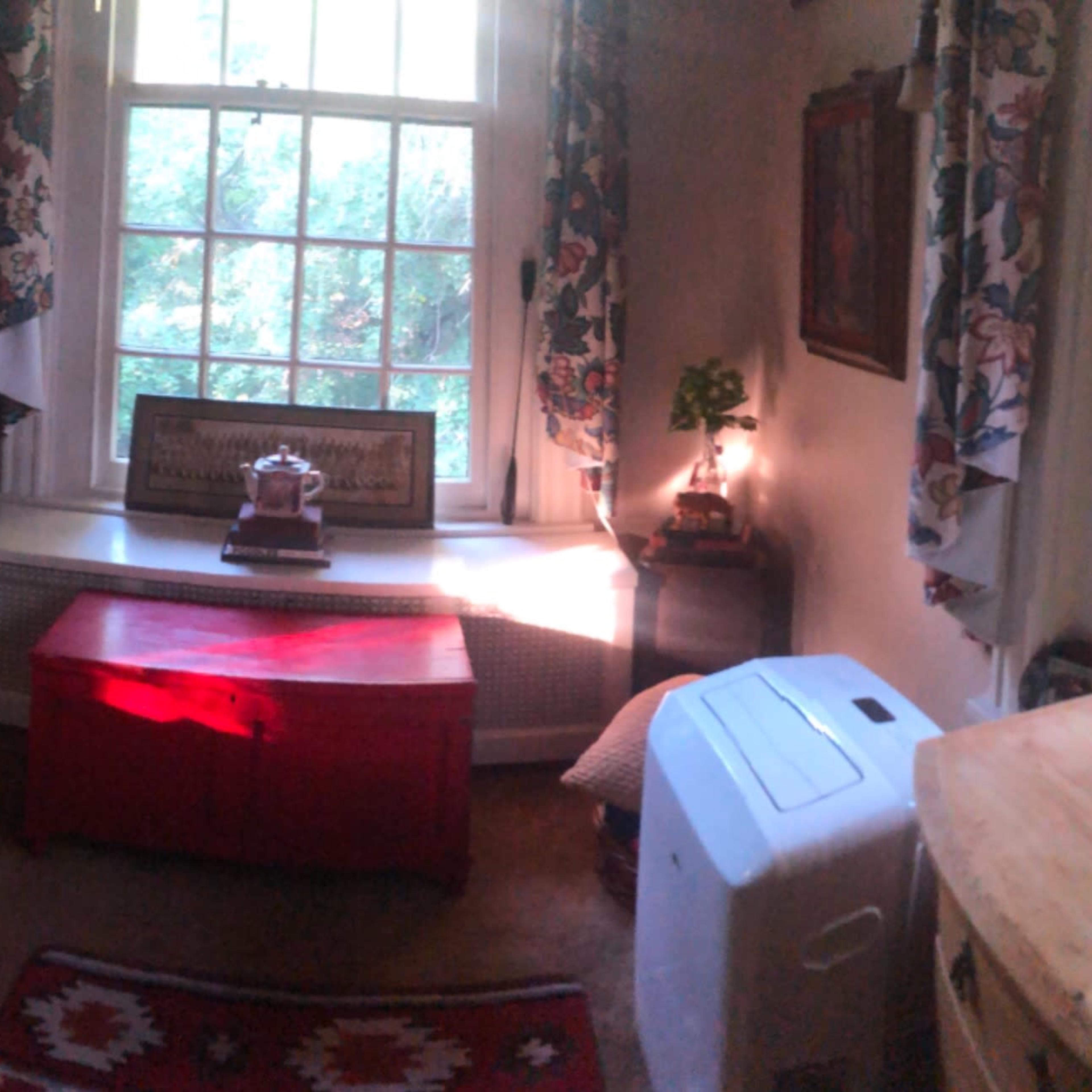A cozy bedroom with a bed, a fan, a desk, a small red table, and windows allowing natural light to enter.