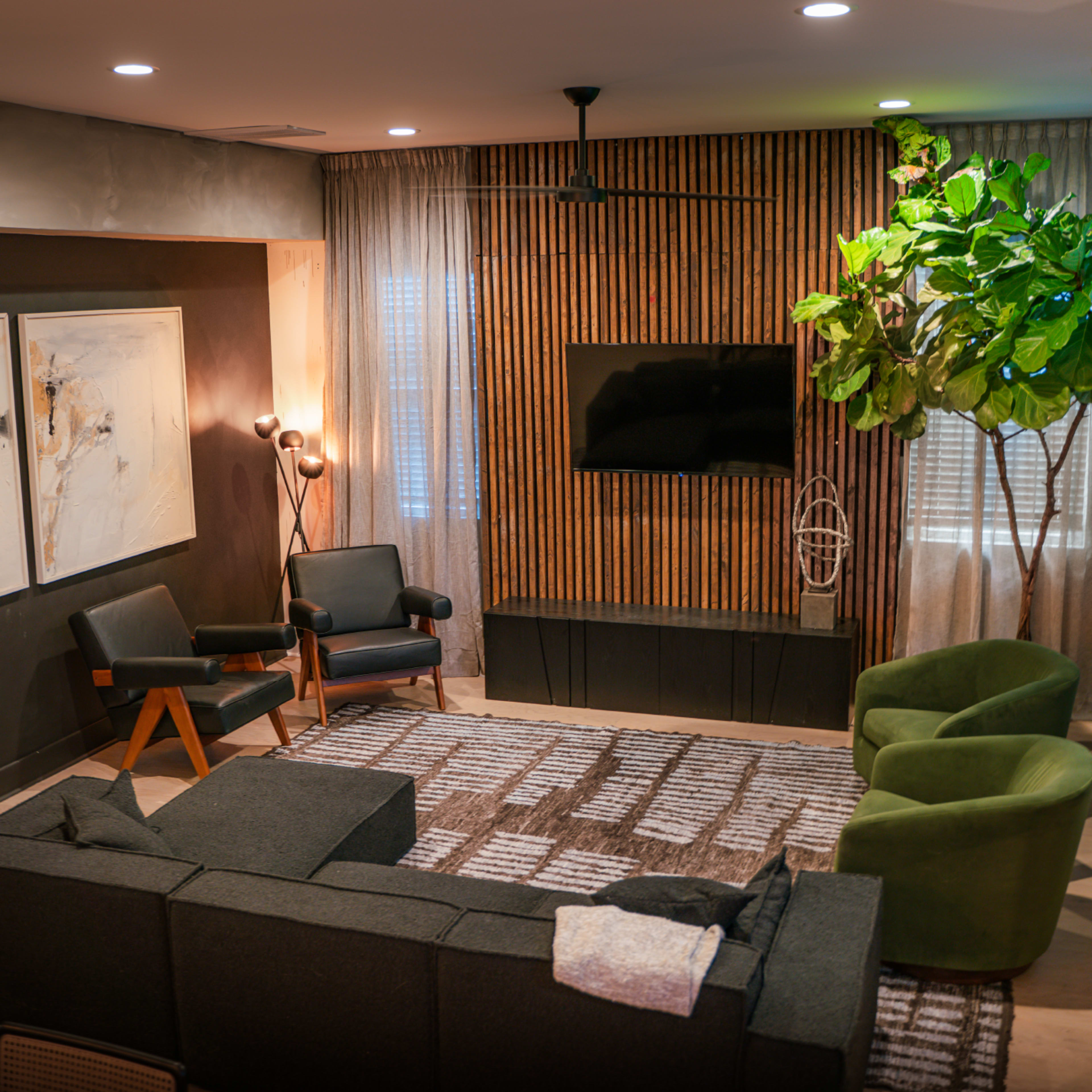 A modern living room featuring a gray sectional sofa, two black armchairs, a green lounge chair, a TV mounted on a wooden panel, and large curtains.