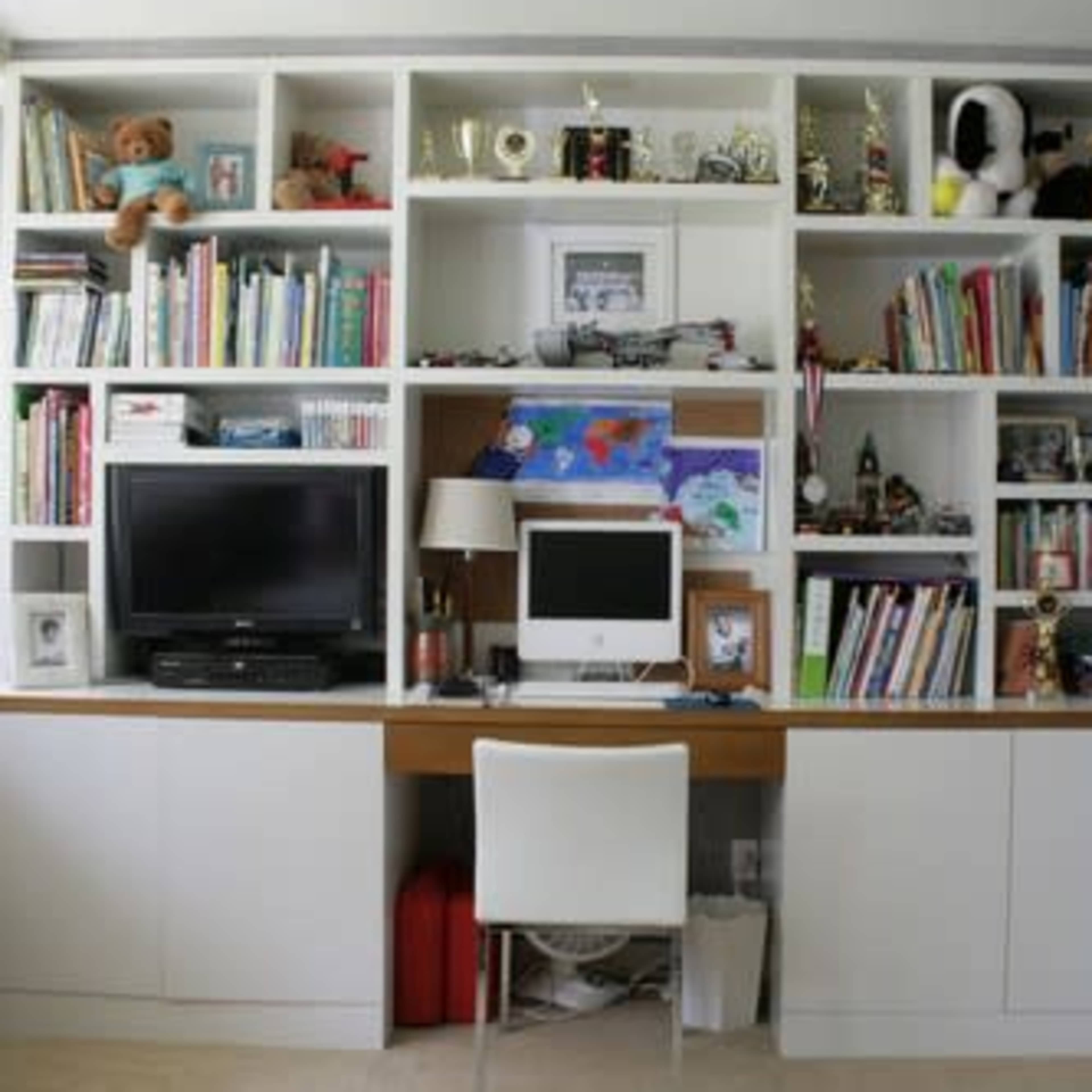 A white shelving unit filled with books, toys, and trophies, with a desk area featuring a television, a computer, and a chair.