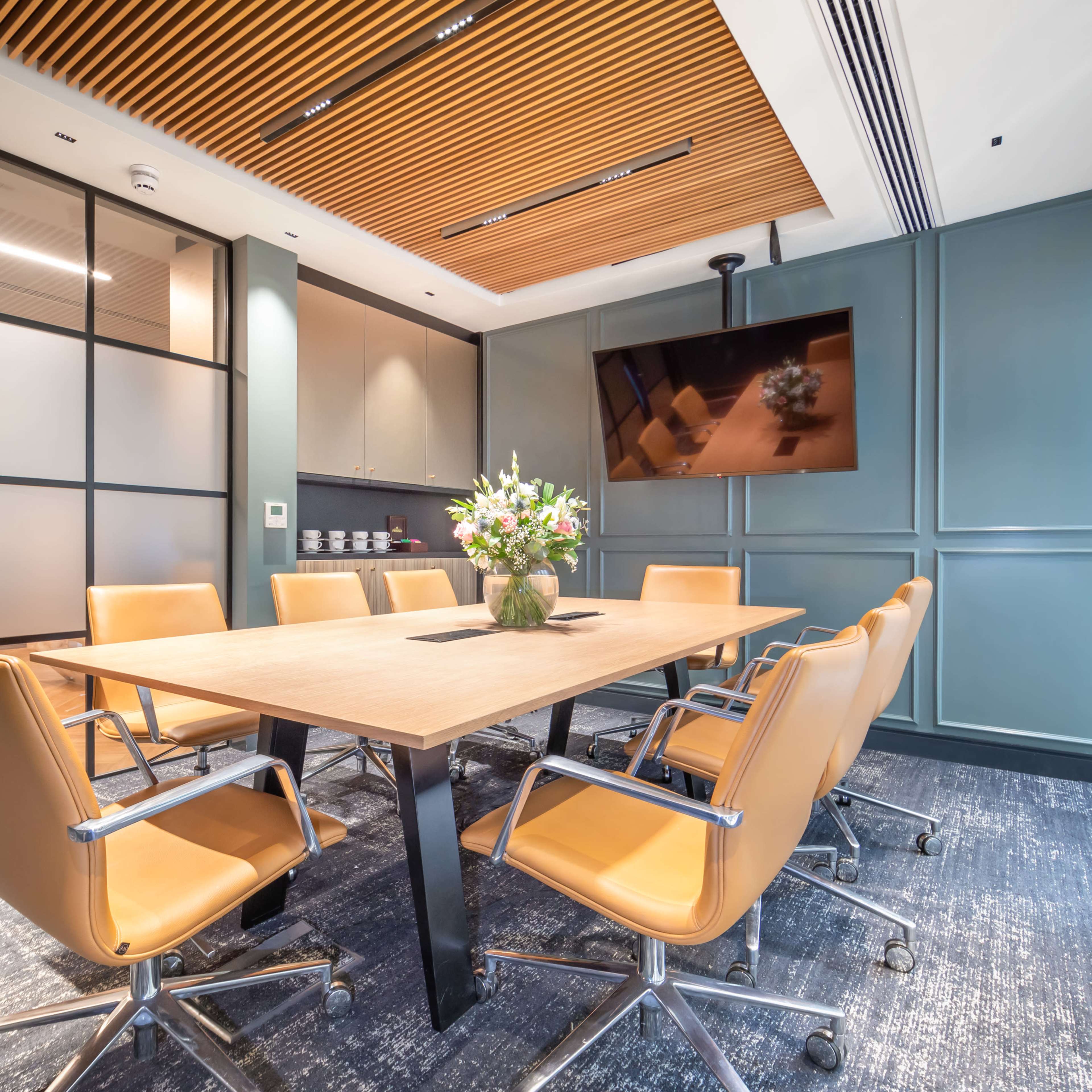 A modern conference room features a large wooden table surrounded by leather chairs, with a wall-mounted television and a floral centerpiece.