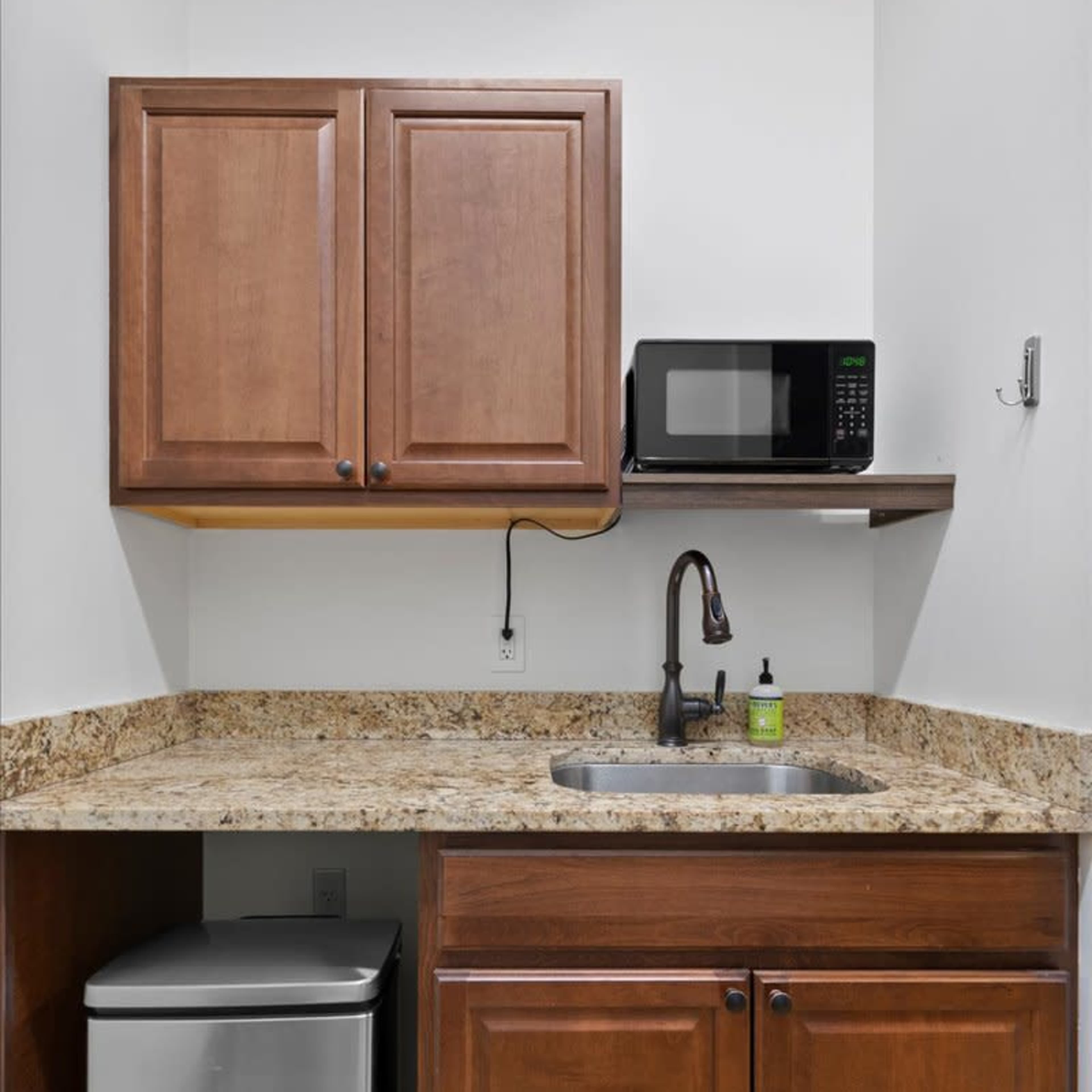 A laundry area with wooden cabinets above a countertop, a sink, a microwave, and a trash can.
