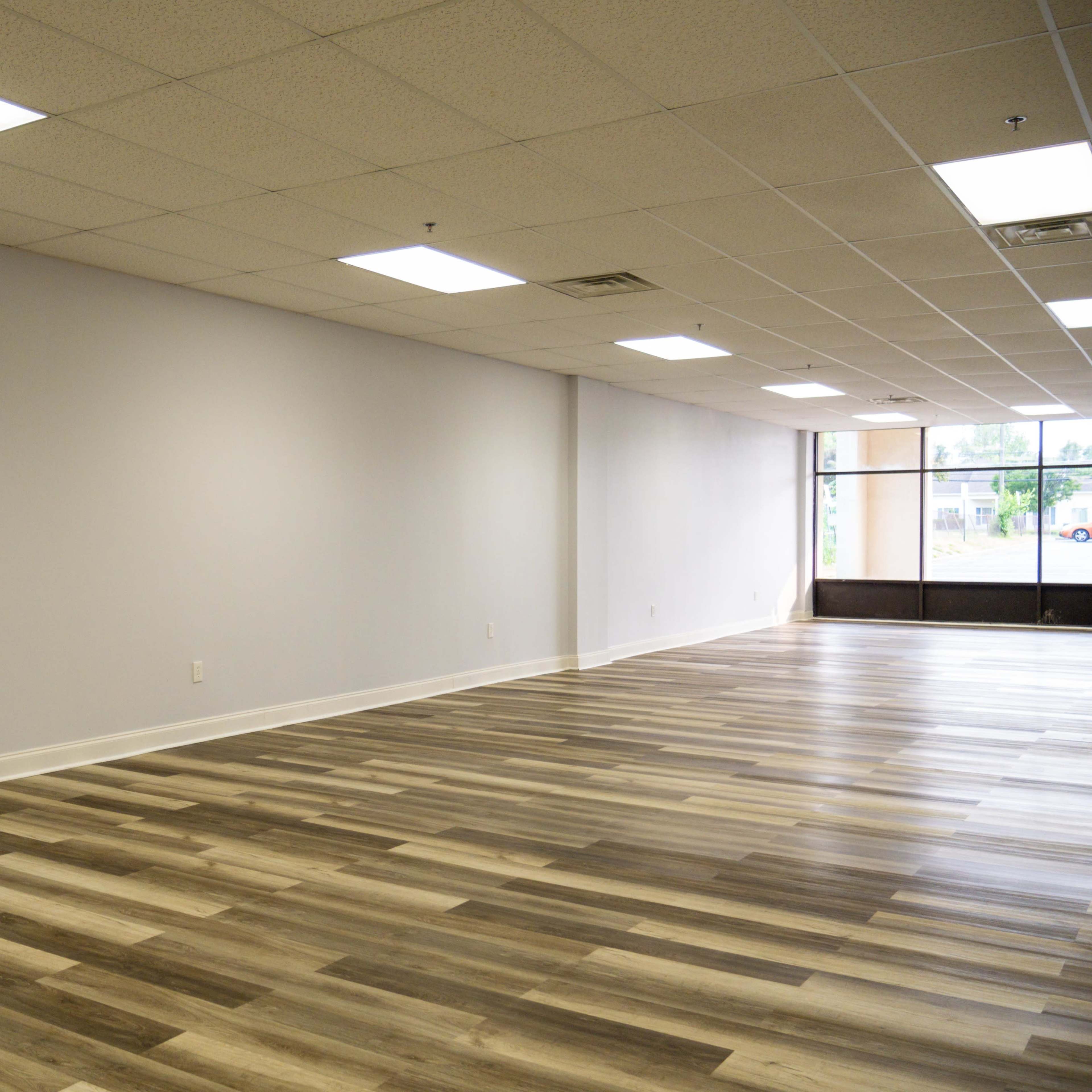 An empty commercial space features light wood flooring and plain white walls under bright ceiling lights.