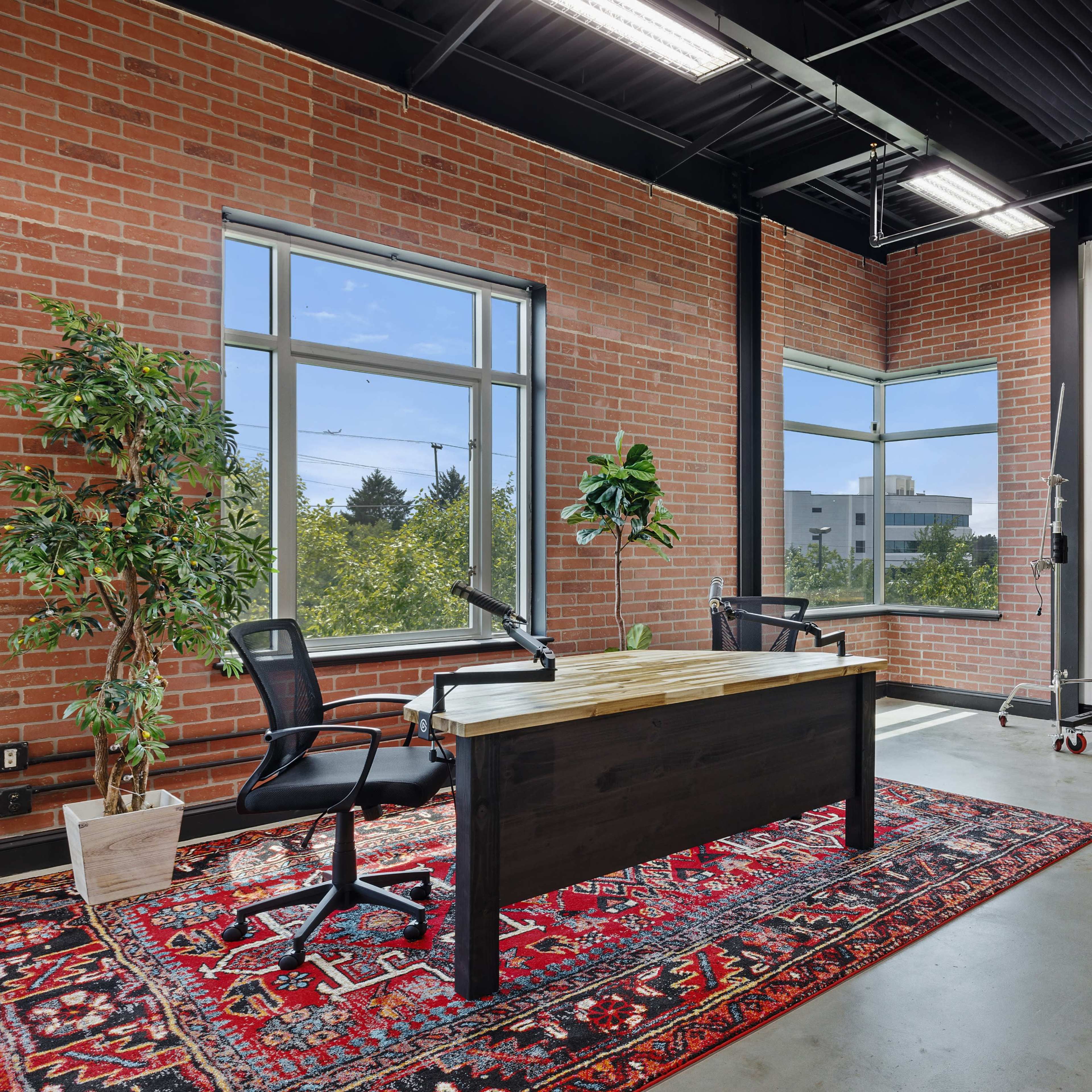 A modern office space featuring a wooden desk, two black chairs, large windows with views, and a wooden partition against a brick wall.