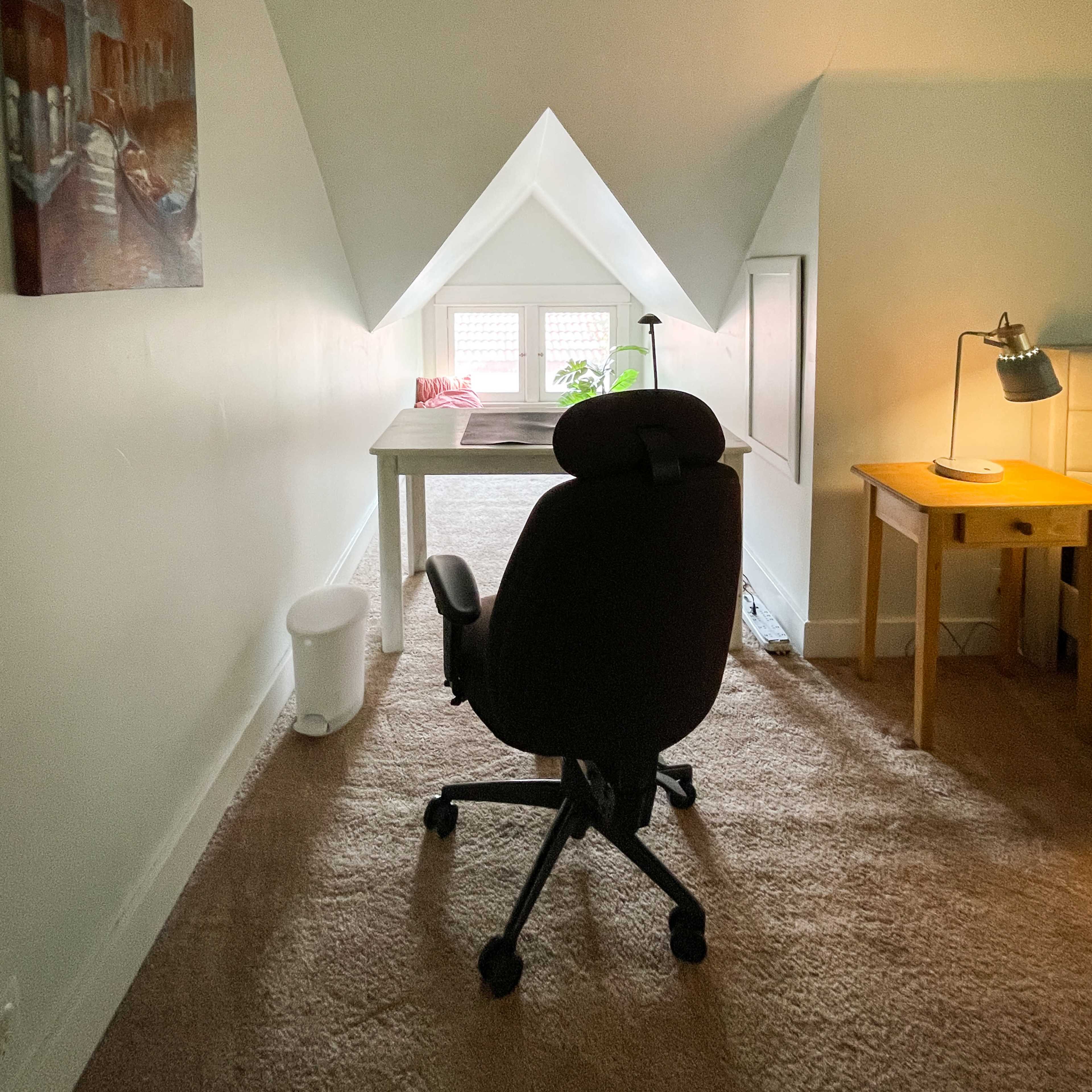 A swivel chair is positioned facing a desk near a window in a cozy, sloped-roof attic space.