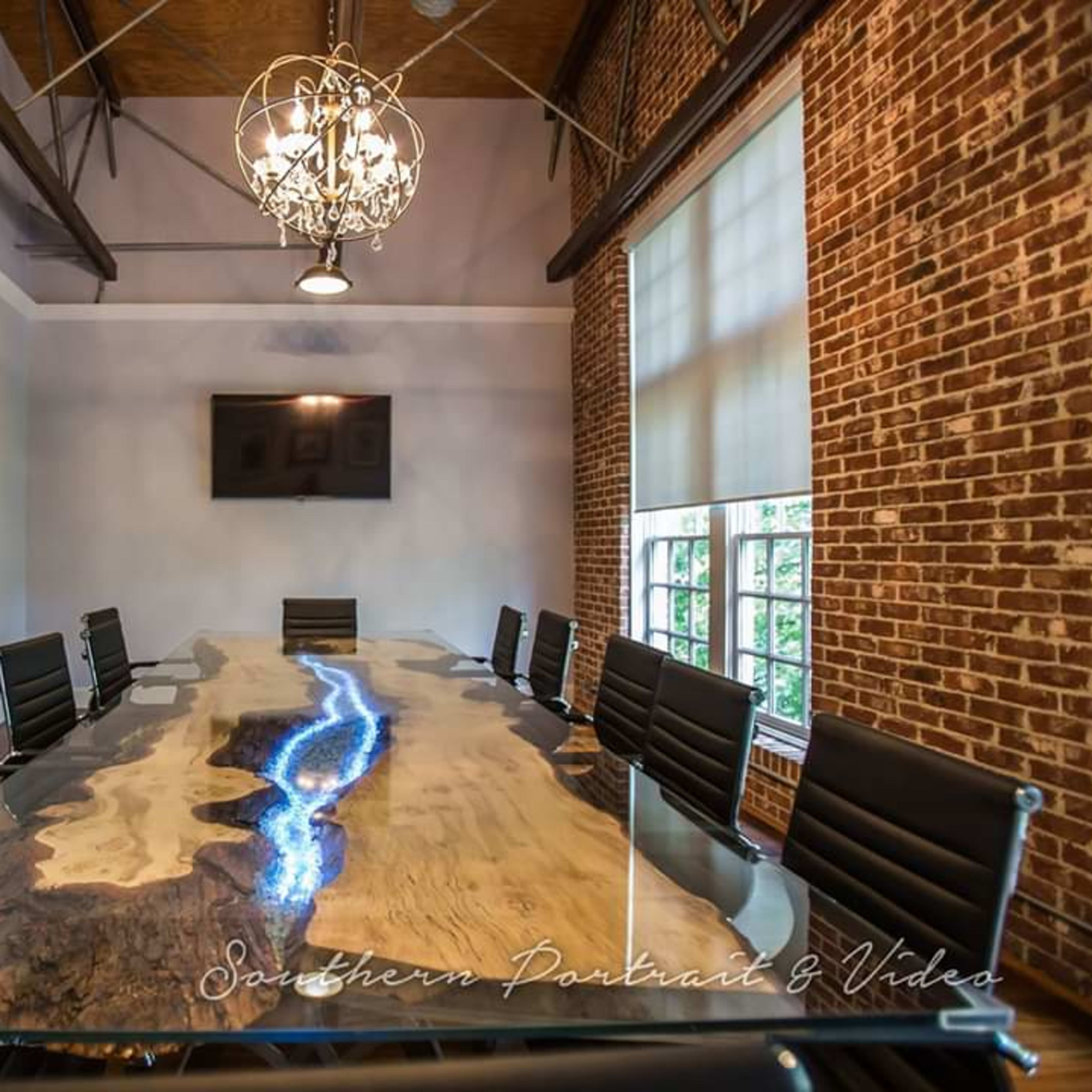 A modern conference room features a long wooden table with a striking blue resin inlay, surrounded by black leather chairs, with brick walls and a chandelier overhead.