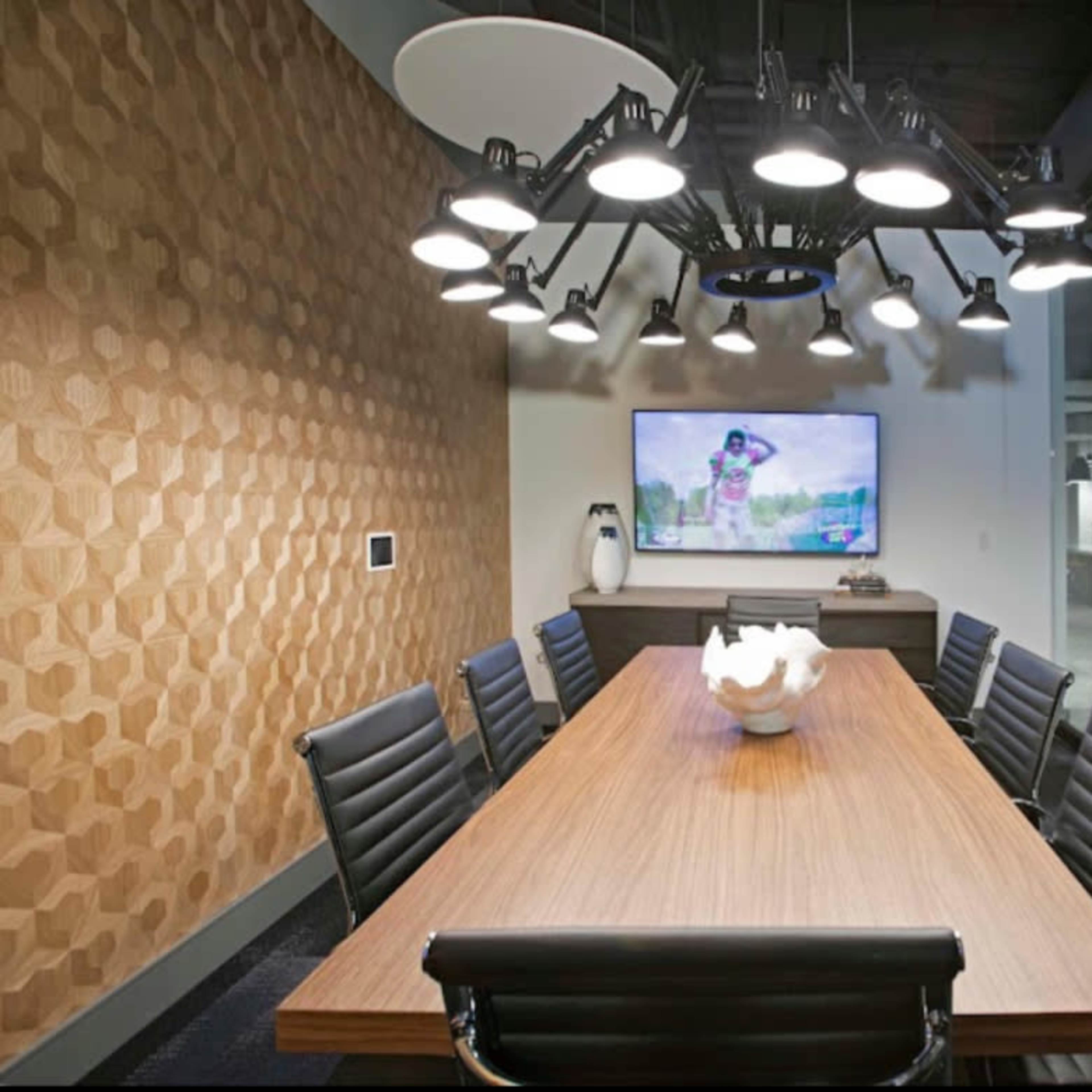 A conference room features a long wooden table surrounded by black chairs, a large screen displaying a presentation, and a modern overhead light fixture.