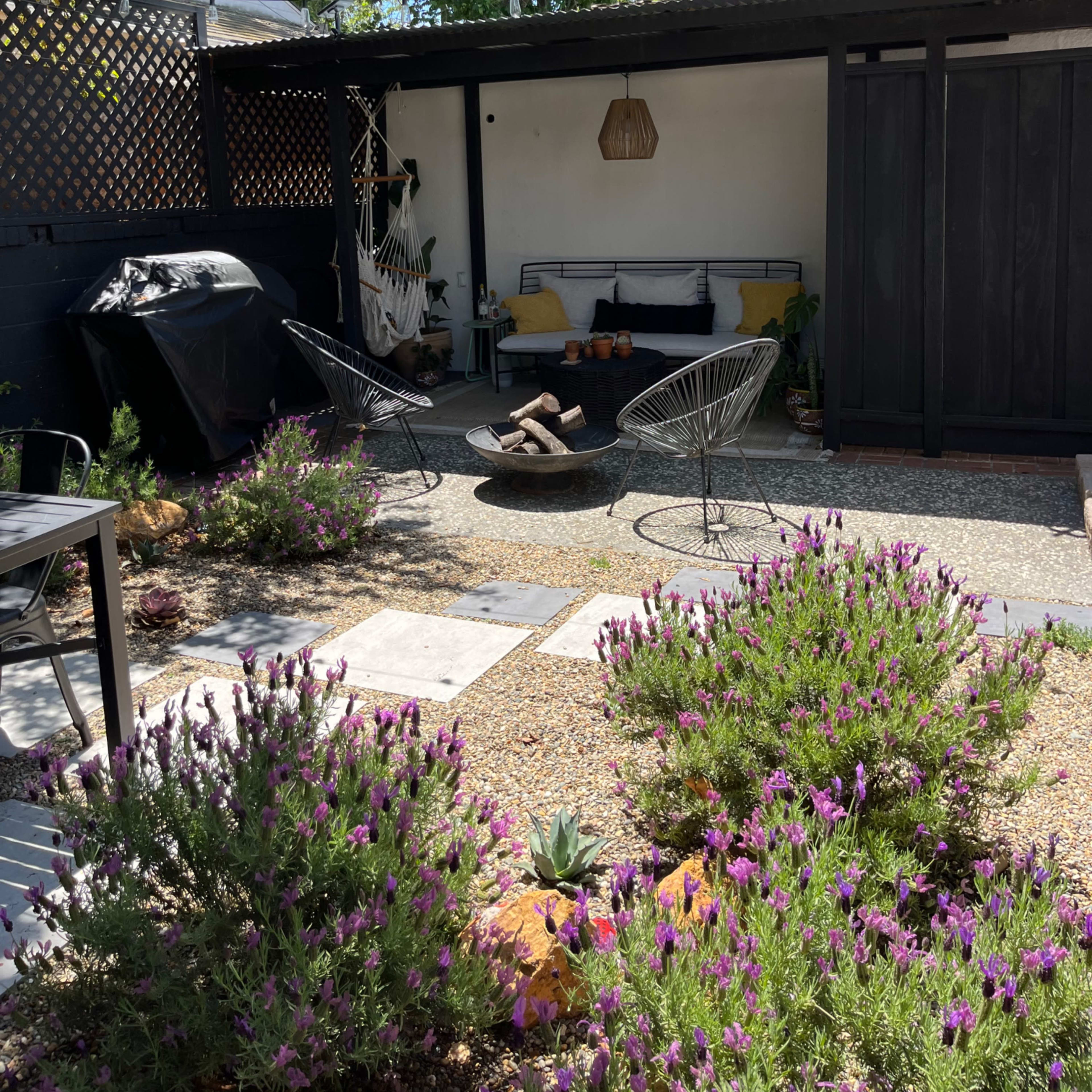 A cozy outdoor seating area features two chairs around a fire pit, surrounded by lavender plants and string lights overhead.
