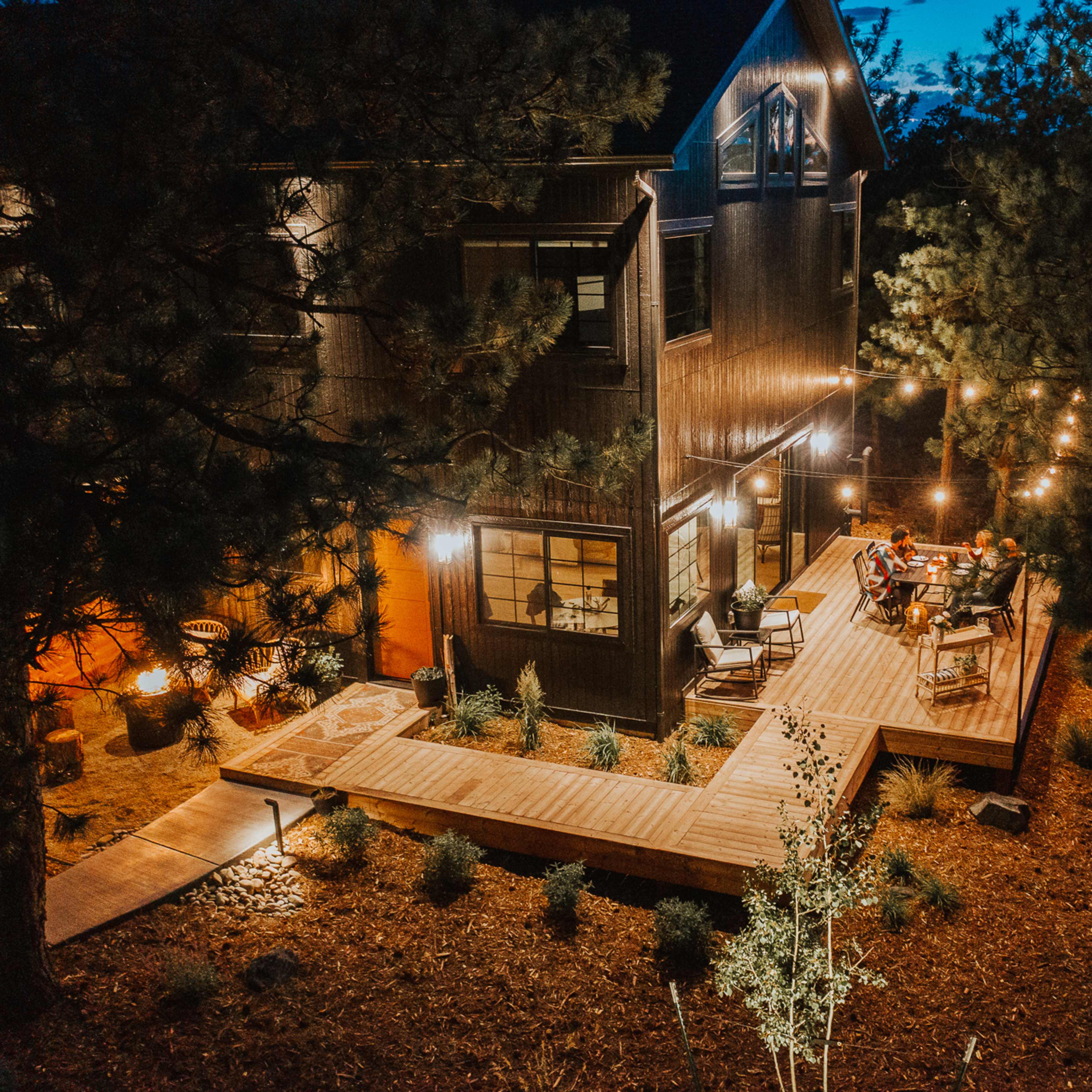 A wooden deck with string lights is set up in front of a large house surrounded by trees, where a group of people is seated, enjoying the evening.