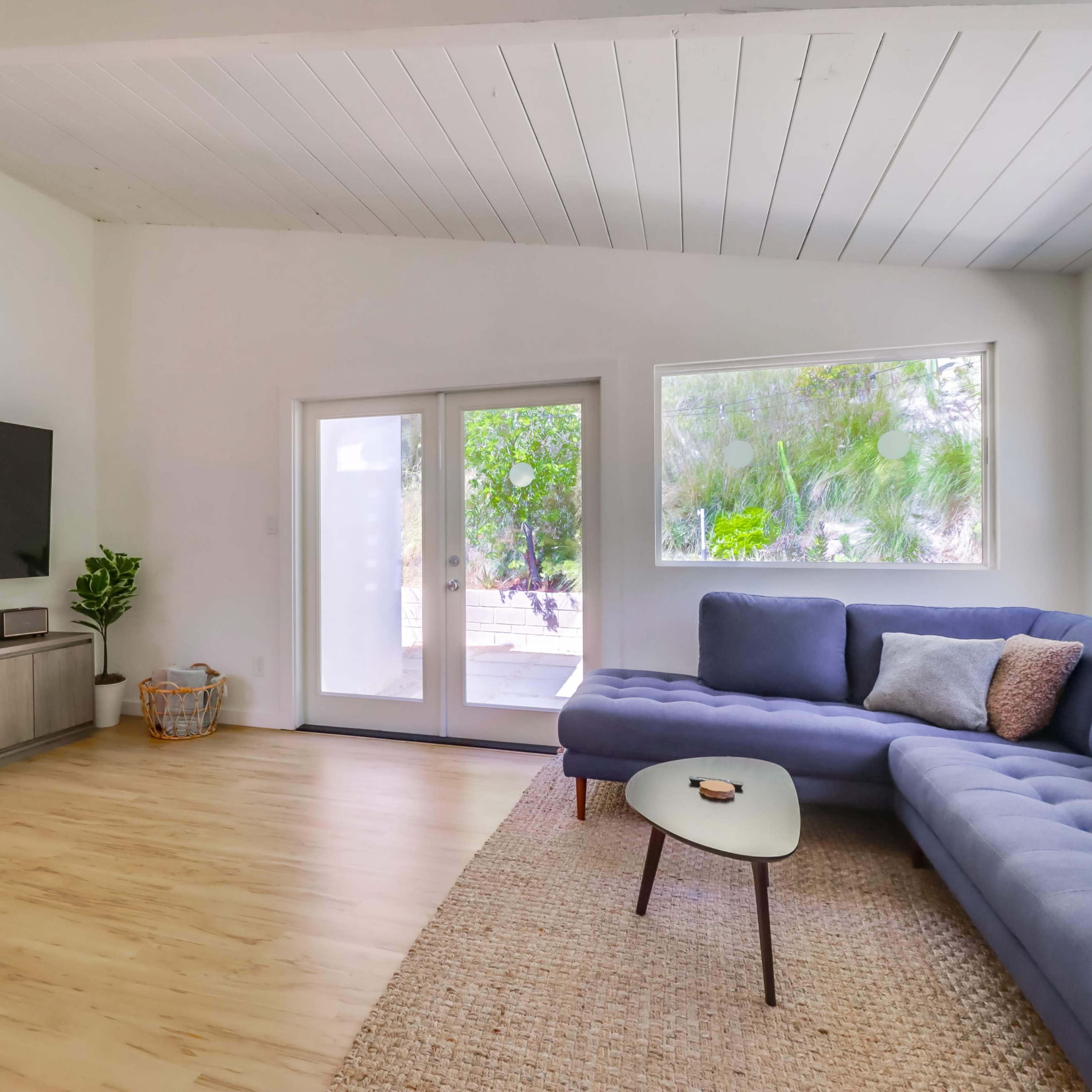 A spacious living room features a blue sectional sofa, a modern coffee table, a television mounted on the wall, and large windows that overlook greenery outside.