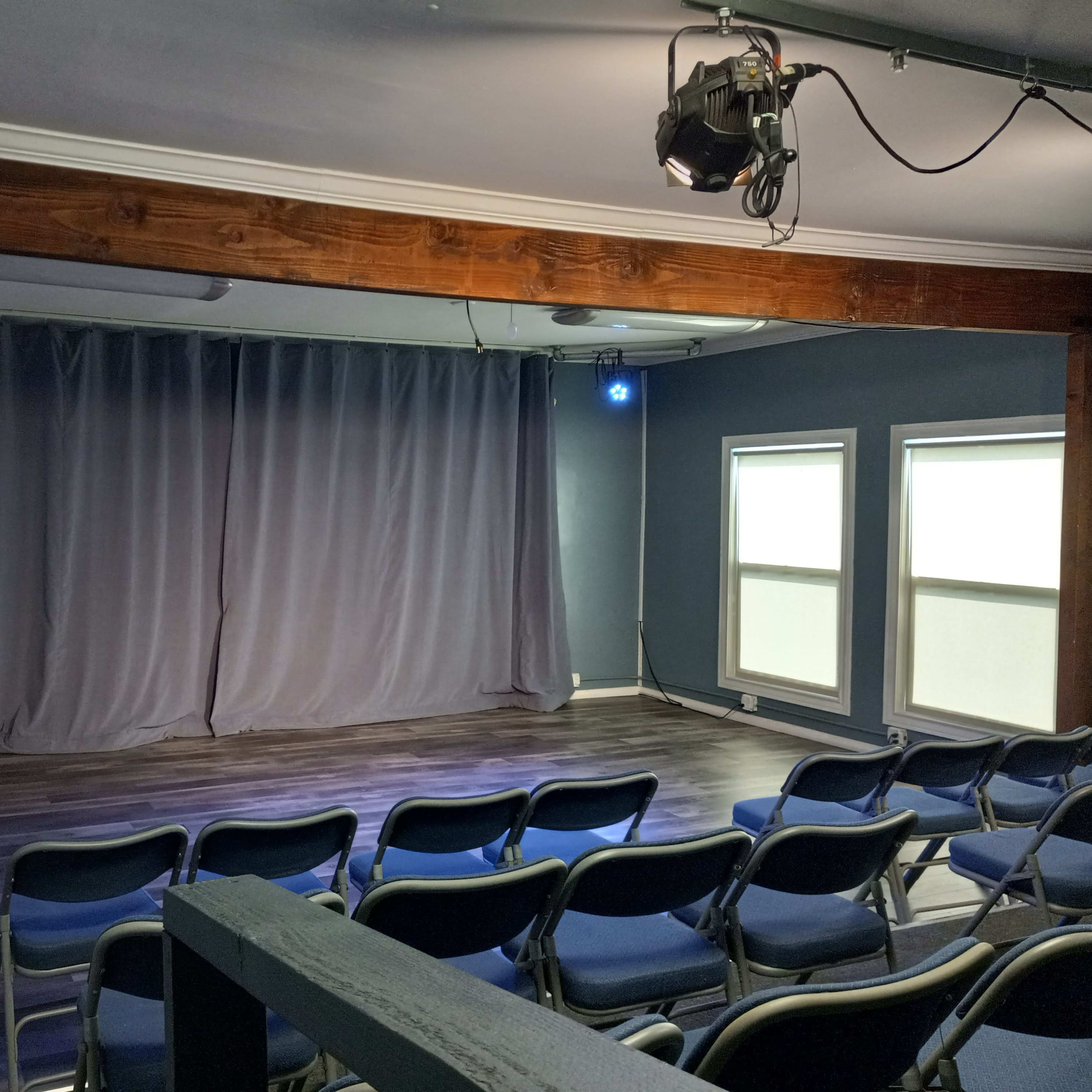 A small performance space with a gray curtain, blue stage lights, and rows of blue chairs facing the stage.
