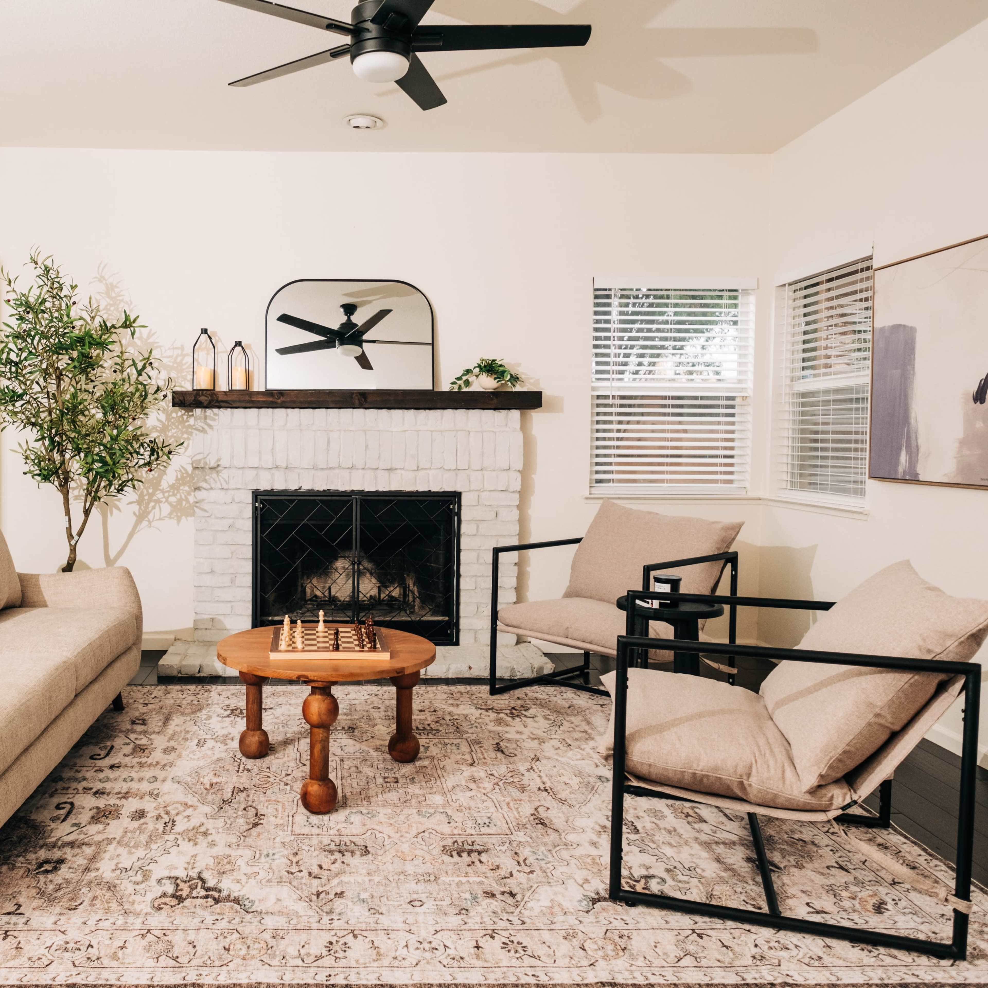 A living room features a beige sofa, two black framed chairs, a wooden coffee table with a chess set, and a fireplace surrounded by white brick.
