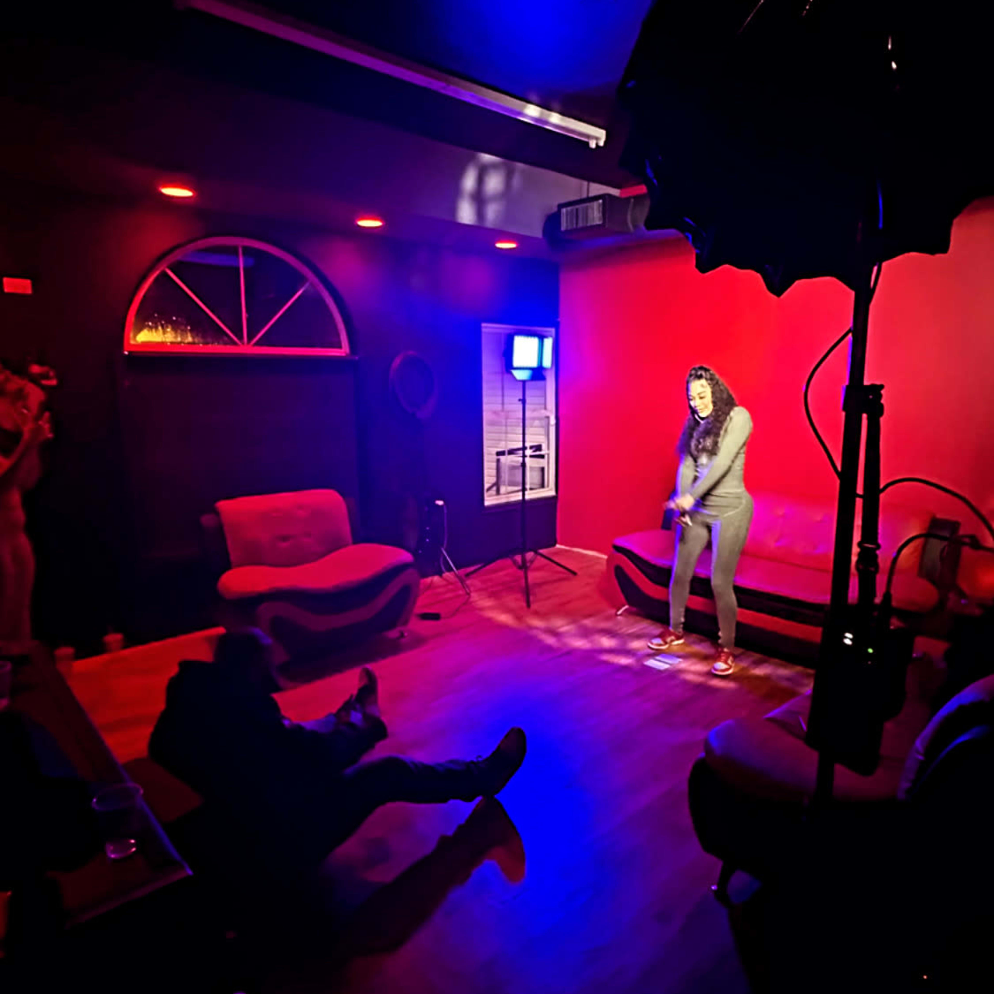 A performer stands on a small stage in a dimly lit room with red walls, while an audience sits on couches, observing the act.