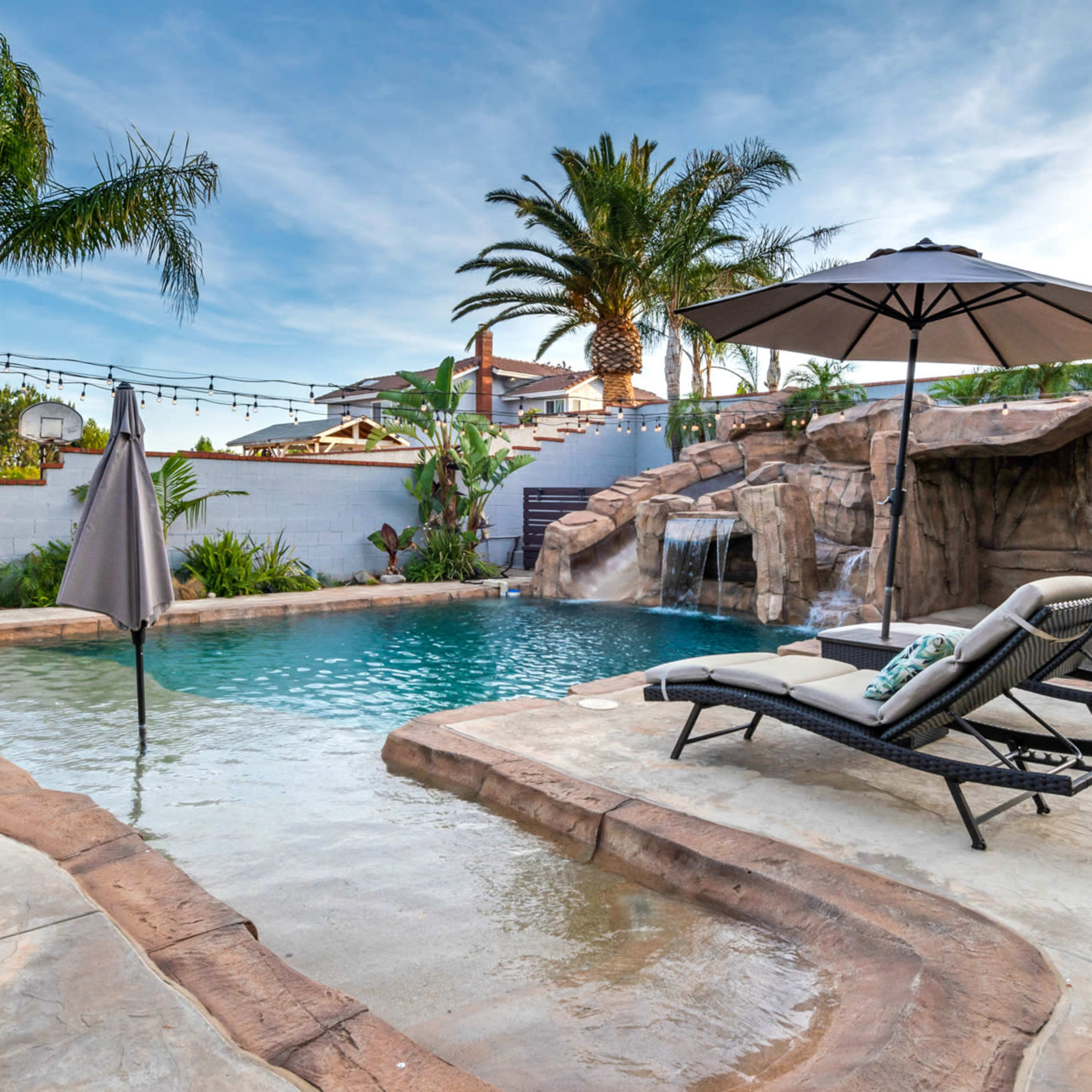 A backyard pool with a rocky waterfall, surrounded by palm trees and lounge chairs under an umbrella.