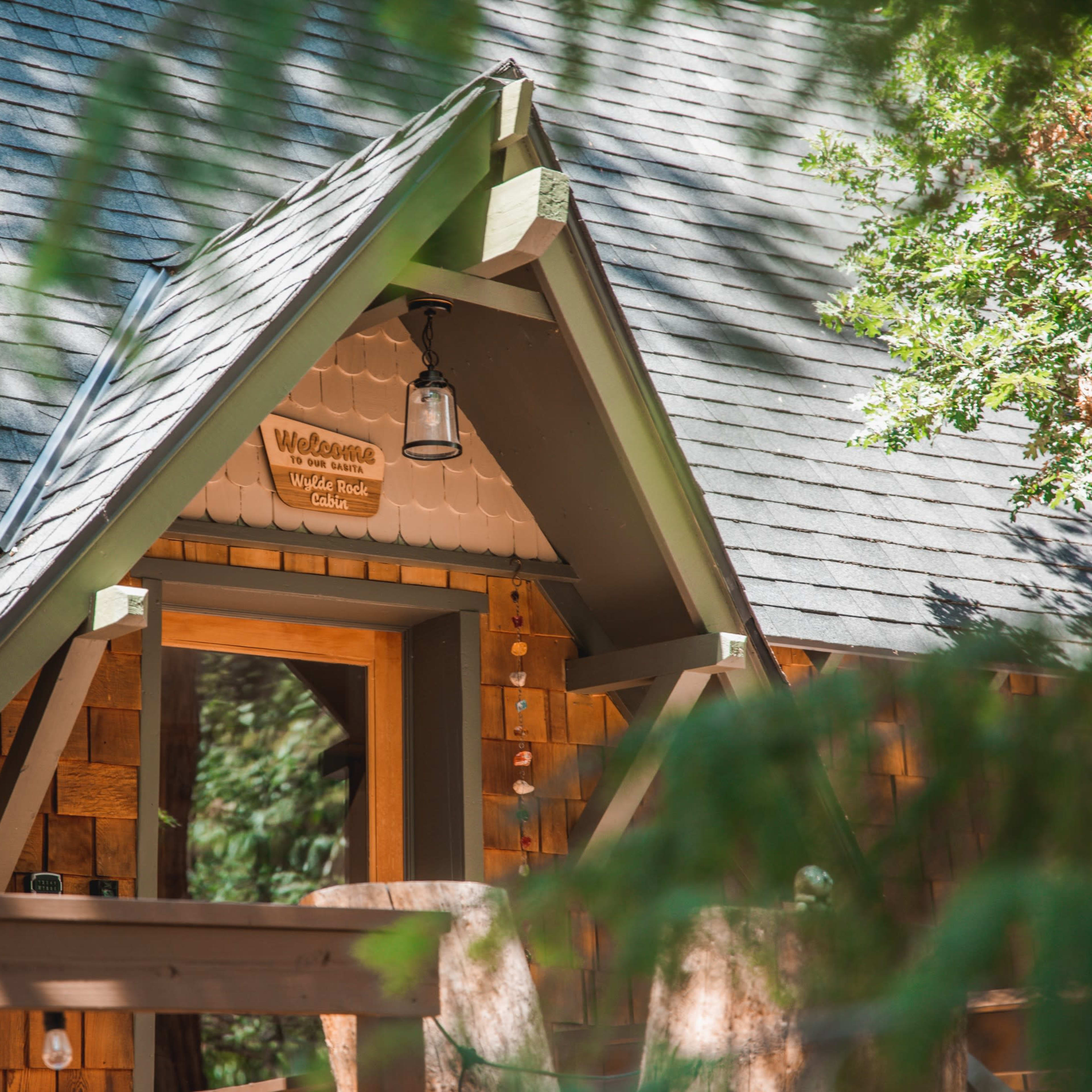 A cabin entrance with a triangular roof, wooden shingles, and a welcome sign above the door, surrounded by trees.