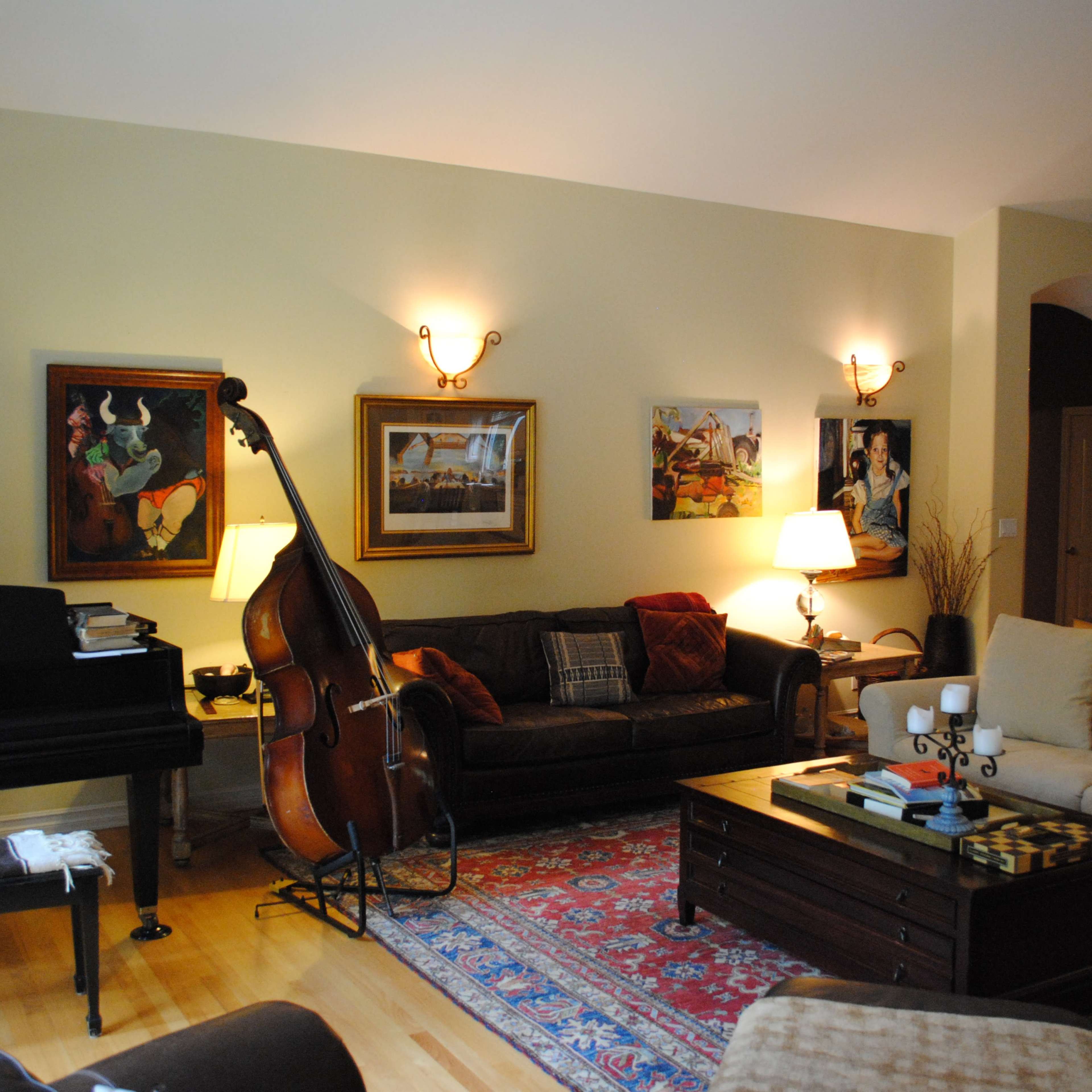 A living room with a piano, a double bass, two sofas, several framed artworks on the walls, and a wooden coffee table in the center.
