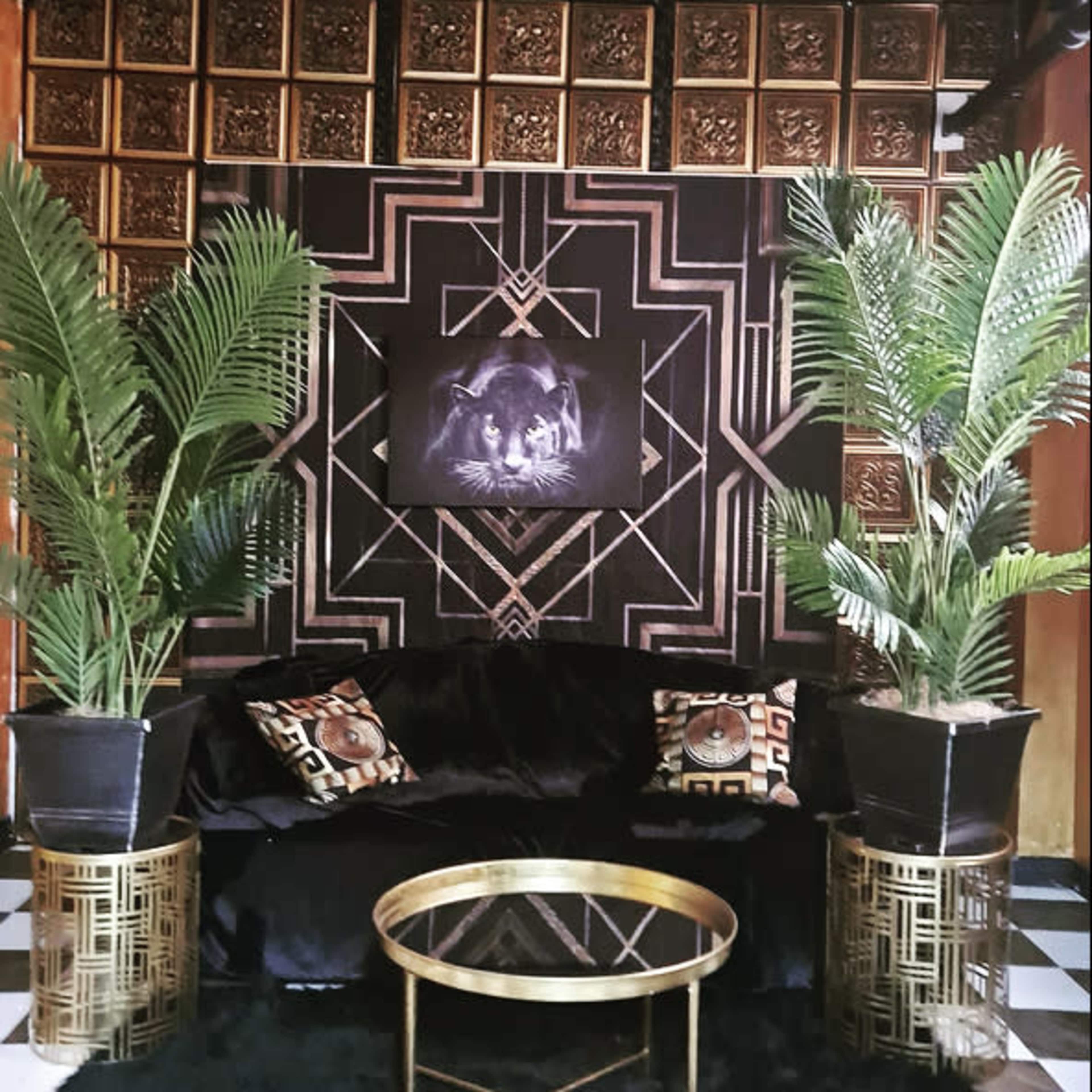 A stylish lounge area featuring a black sofa, gold-accented side tables, a patterned wall behind, and tall potted plants on either side.